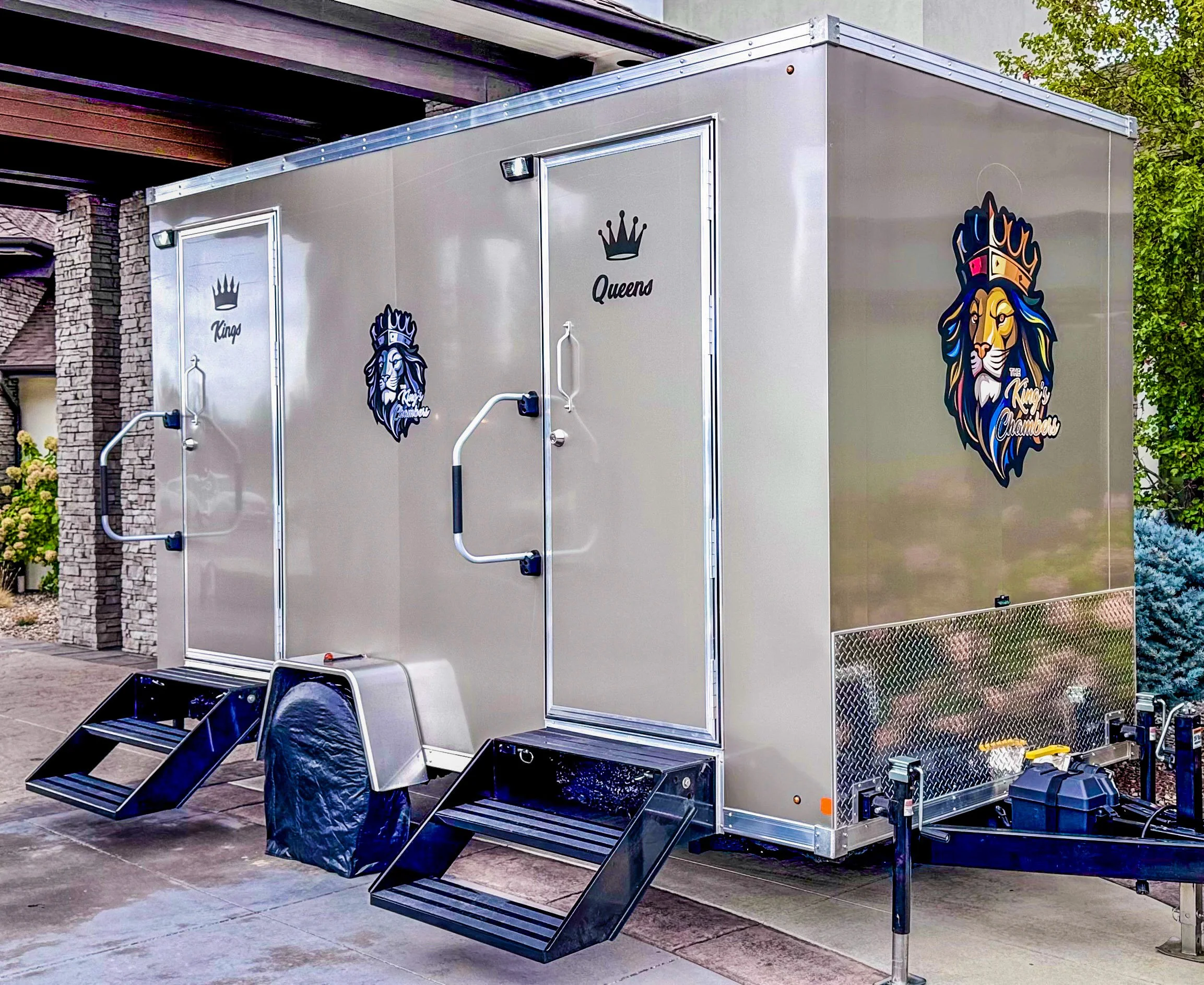 A shiny silver trailer with black stairs leading to the door, decorated with a lion's head logo and crown labeled 'Queen' on the side, parked outdoors.