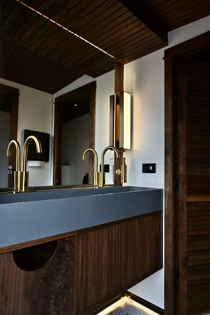 Modern bathroom vanity with gold fixtures, a gray sink, wood accents, and a mirror with vertical lighting.