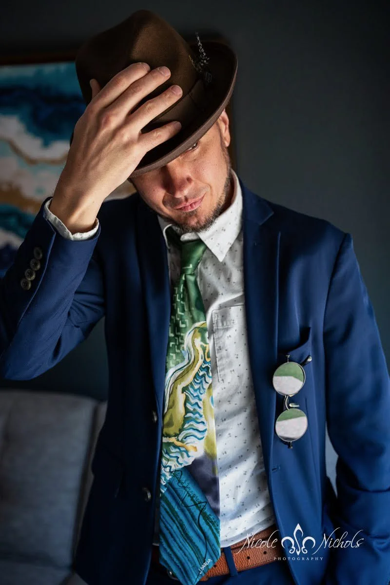A man in a blue suit and white patterned shirt, holding a brown fedora hat, with sunglasses hanging from his pocket, standing indoors against a dark background.