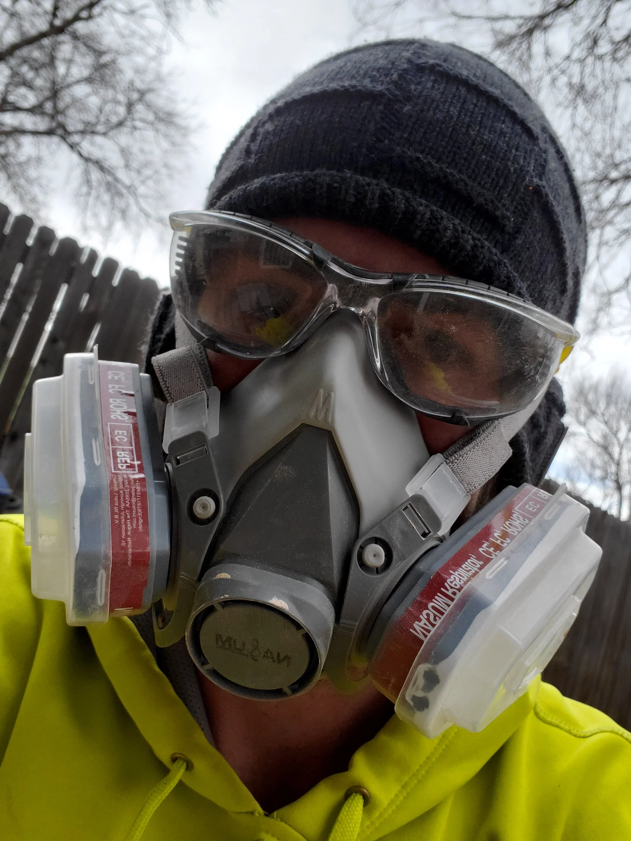 A person wearing a black knit beanie, safety goggles, and a yellow high-visibility jacket holding a medical oxygen tank indoors, with a background of bare tree branches and a cloudy sky.