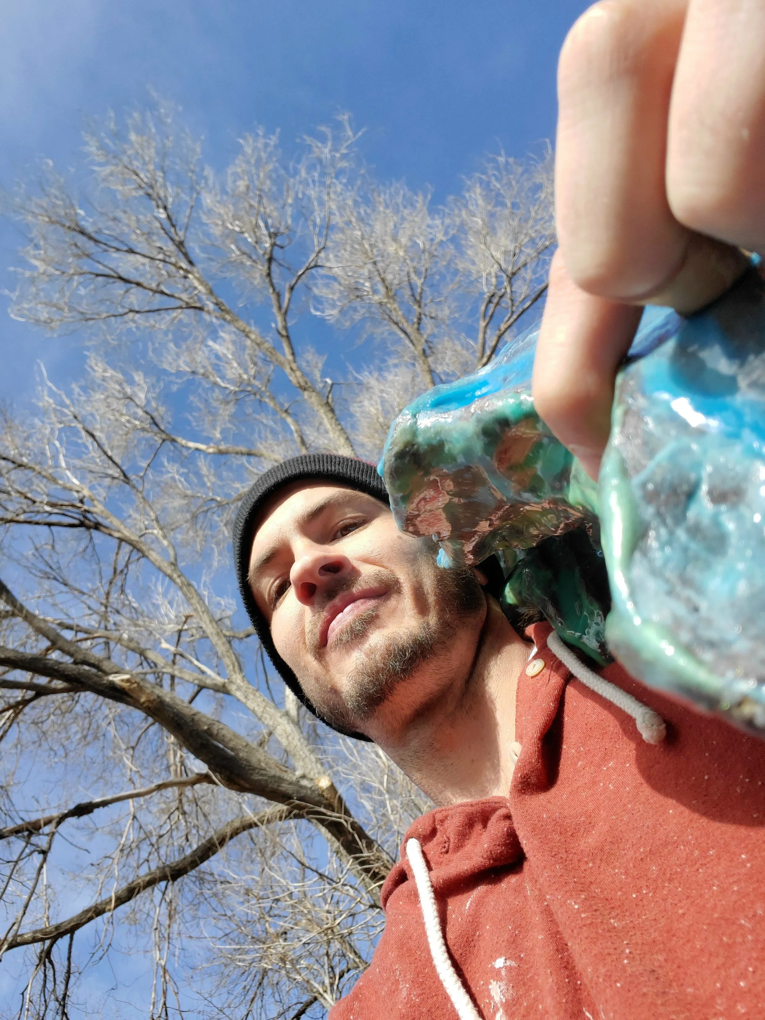 A man wearing a red hoodie and black beanie taking a selfie outdoors. The background features a clear blue sky and leafless trees, indicating it is winter.