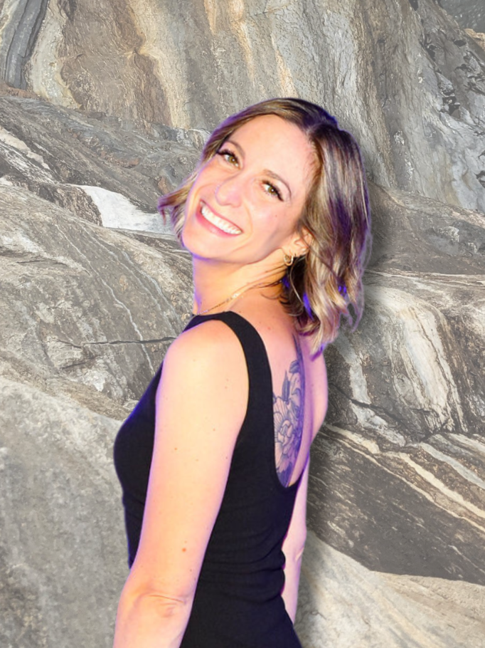 A woman with shoulder-length wavy hair, smiling, in front of a rocky background, wearing a black sleeveless top and gold earrings.