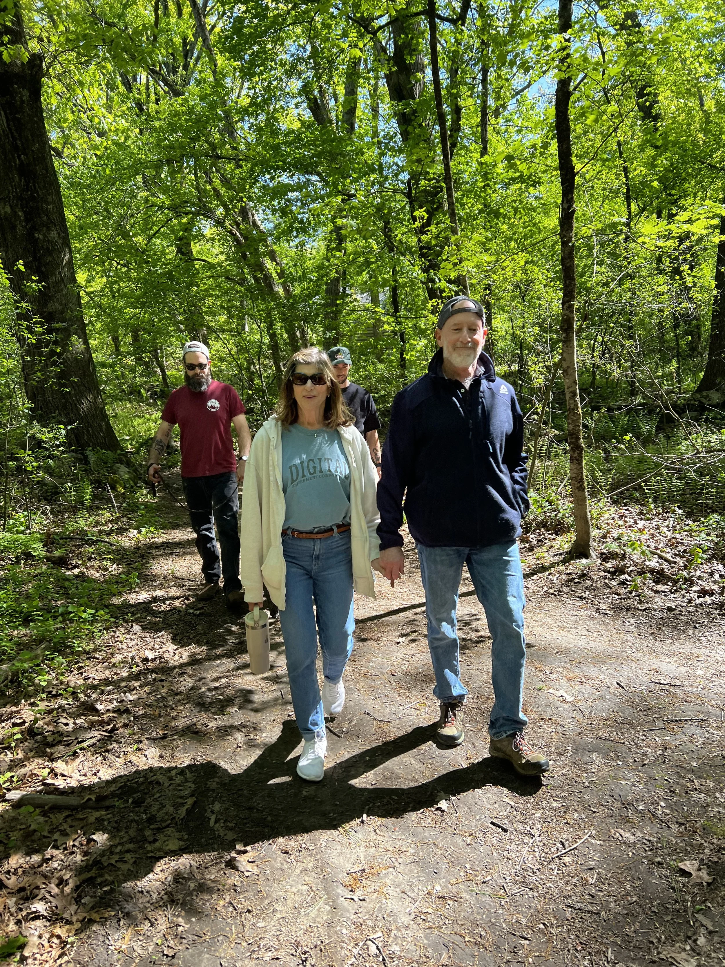 Four people walking on a forest trail surrounded by green trees and sunlight filtering through the leaves. They appear to be on a hike or nature walk.