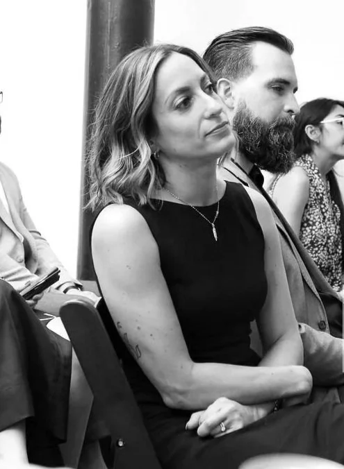 A woman with shoulder-length wavy hair, wearing a sleeveless black dress and a necklace, sitting with her arms crossed, appears attentive during a meeting or event.