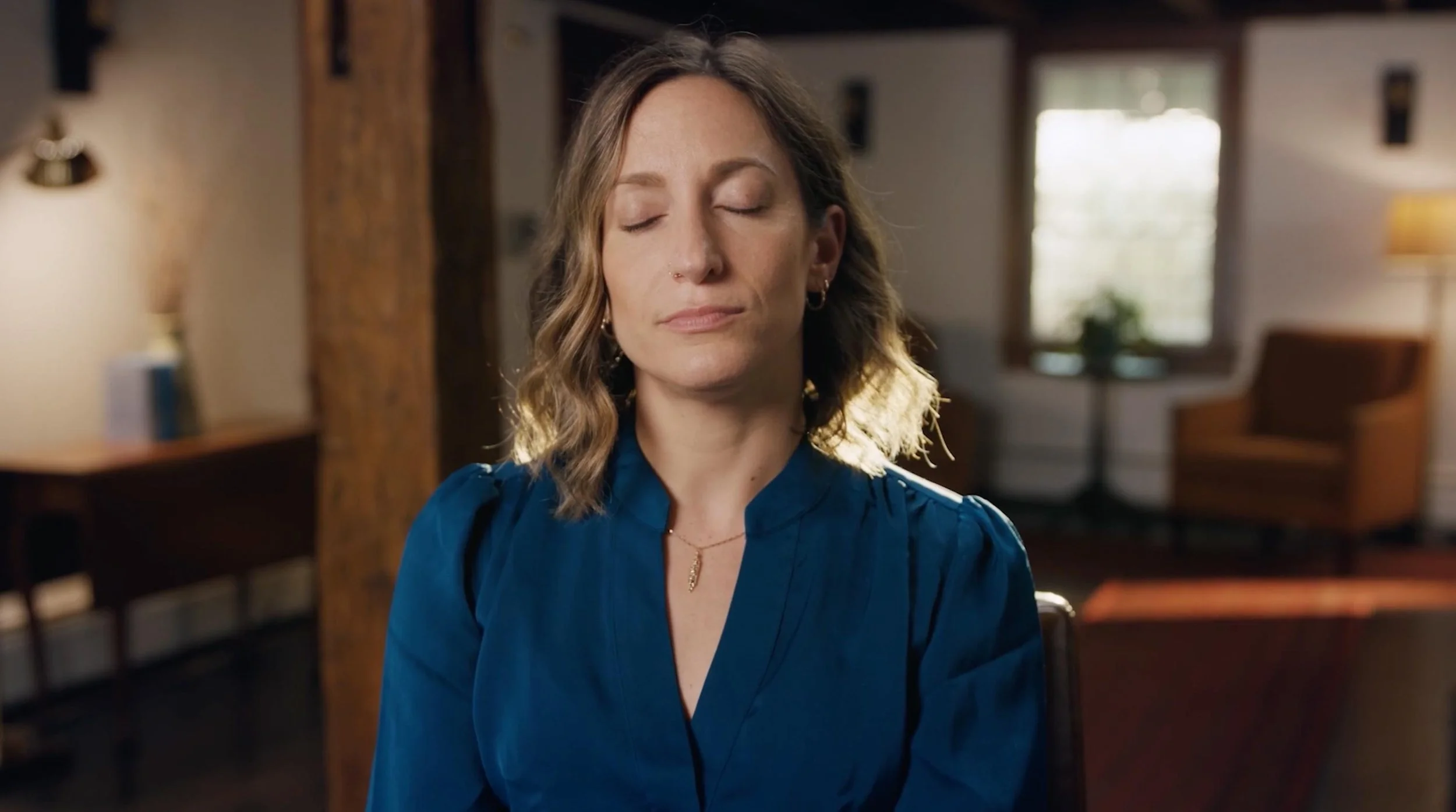 A woman with light skin and wavy, shoulder-length hair wearing a blue blouse with closed eyes, in a cozy room with wooden beams, windows, and soft lighting.