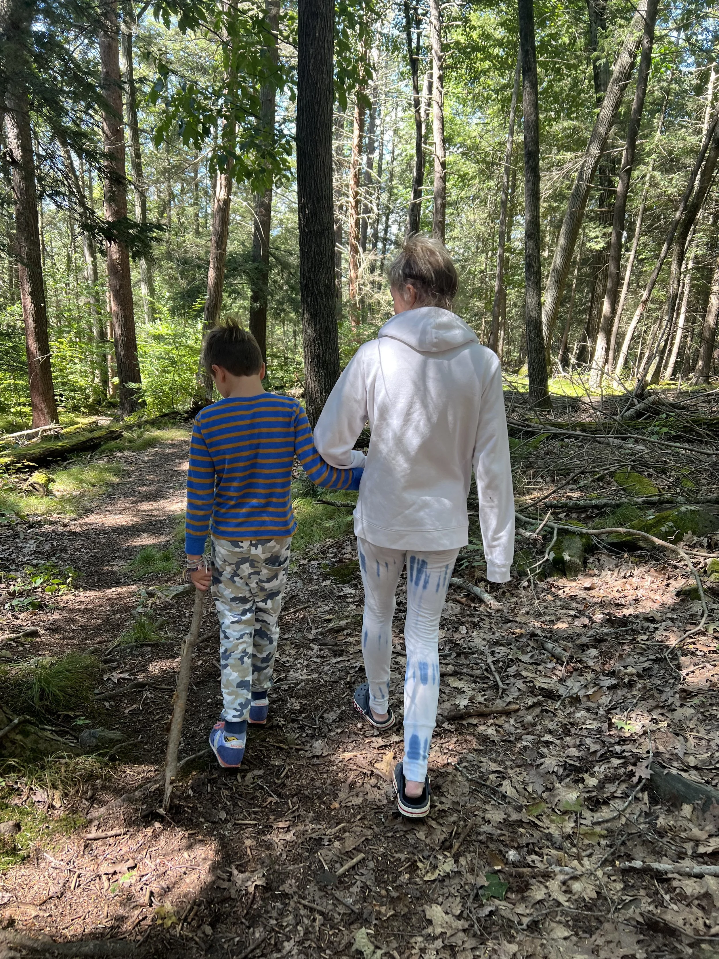 A woman and a boy walking on a forest trail surrounded by tall trees, with sunlight filtering through the leaves.