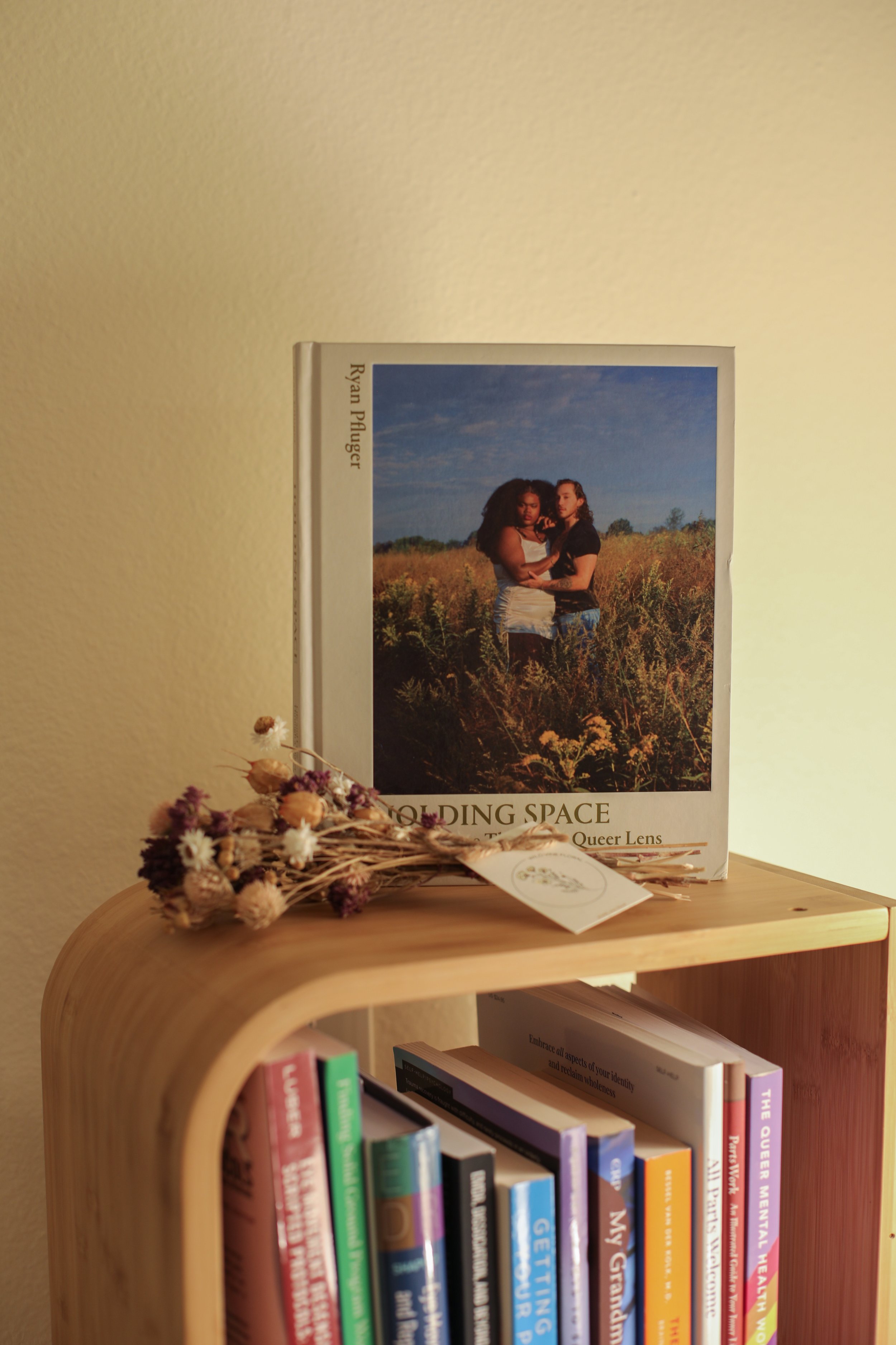 Decorative shelf with therapy books about trauma, EMDR, and parts work, a dried flower arrangement, and a book featuring a queer, interracial couple embracing in a field under a blue sky.