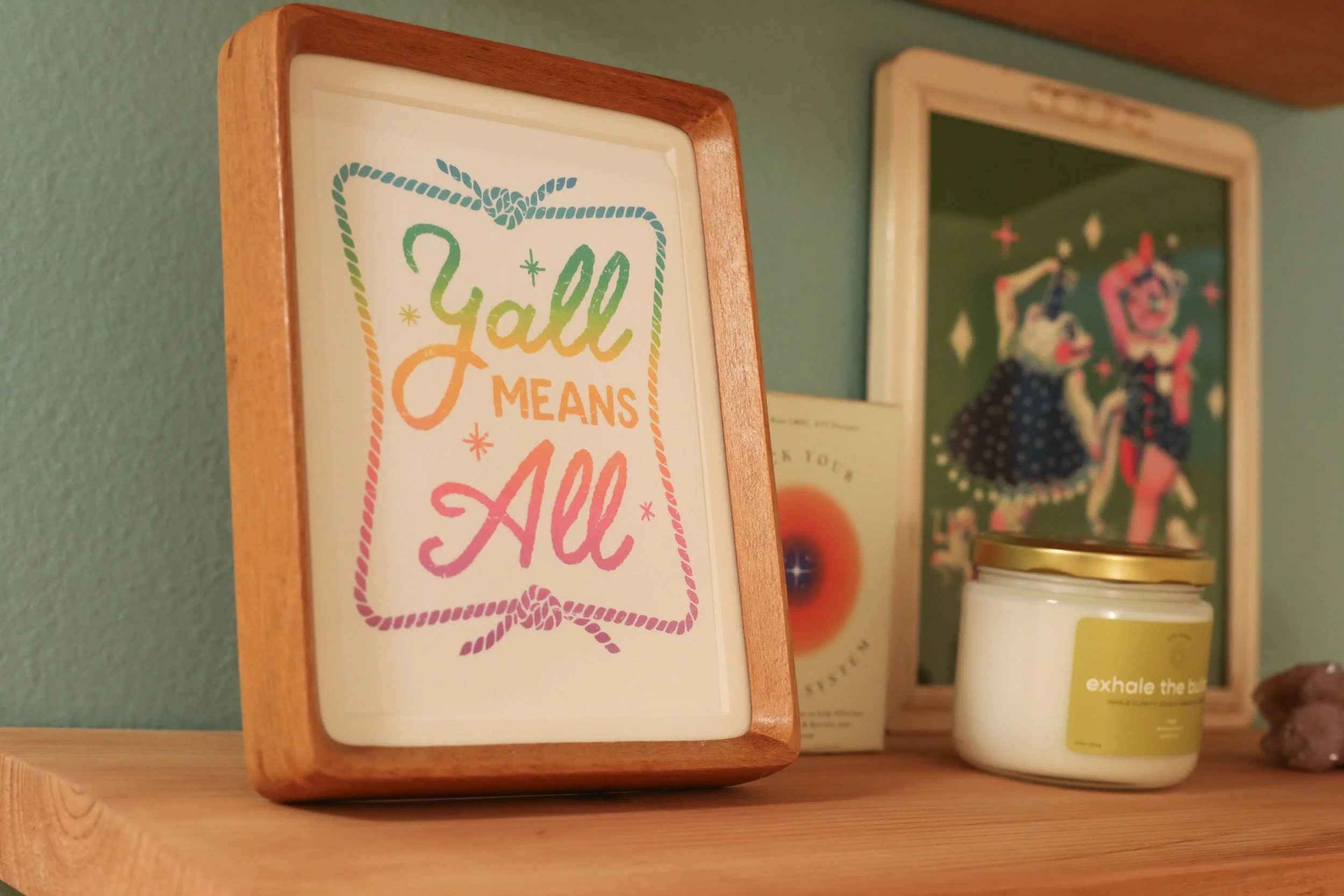 Framed art print with the text 'Y'all Means All' in rainbow cursive, on a wooden shelf alongside various artwork and a candle. 