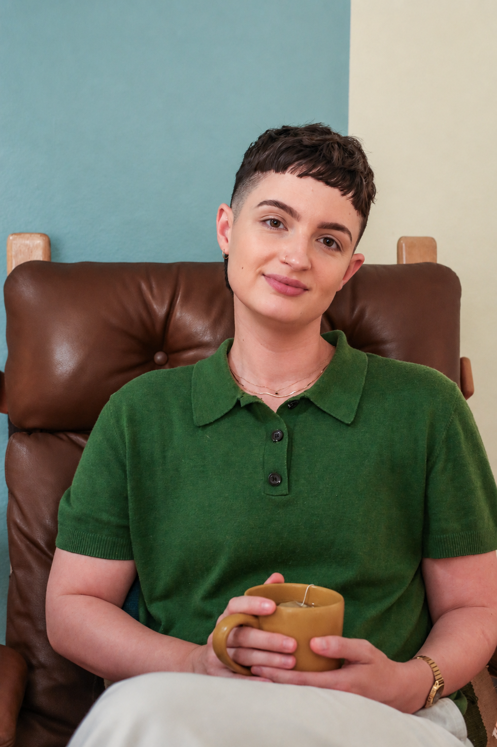 Gay therapist with short dark hair and a warm smile, wearing a green polo shirt, sitting in a brown leather chair against a multicolored wall, holding a yarrow tea mug.