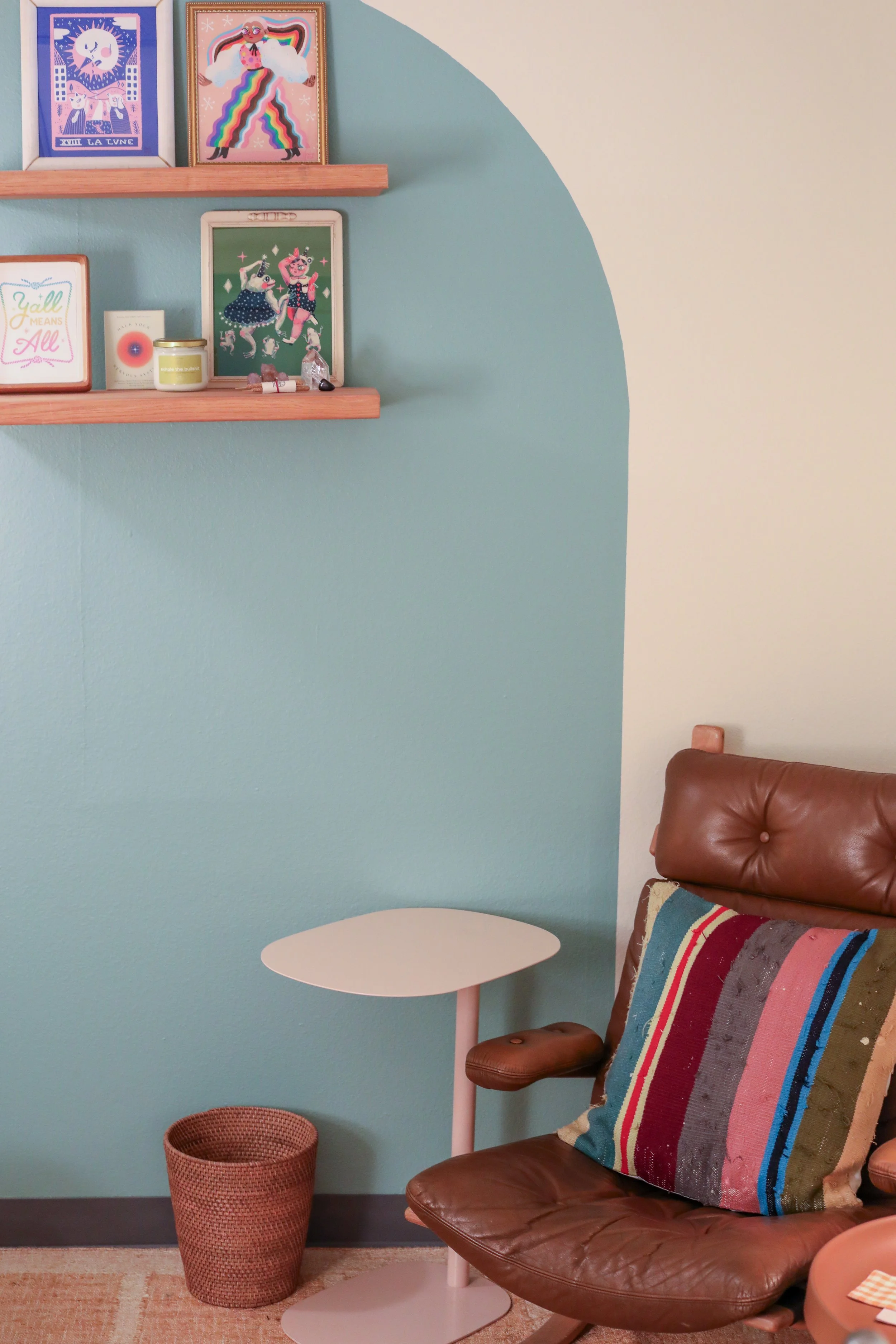 A cozy therapy room corner with sage and cream walls, featuring three colorful framed artworks on a wooden shelf, a pink side table, a brown leather armchair with a multicolored striped pillow, a small woven wastebasket, and a rug on the floor.
