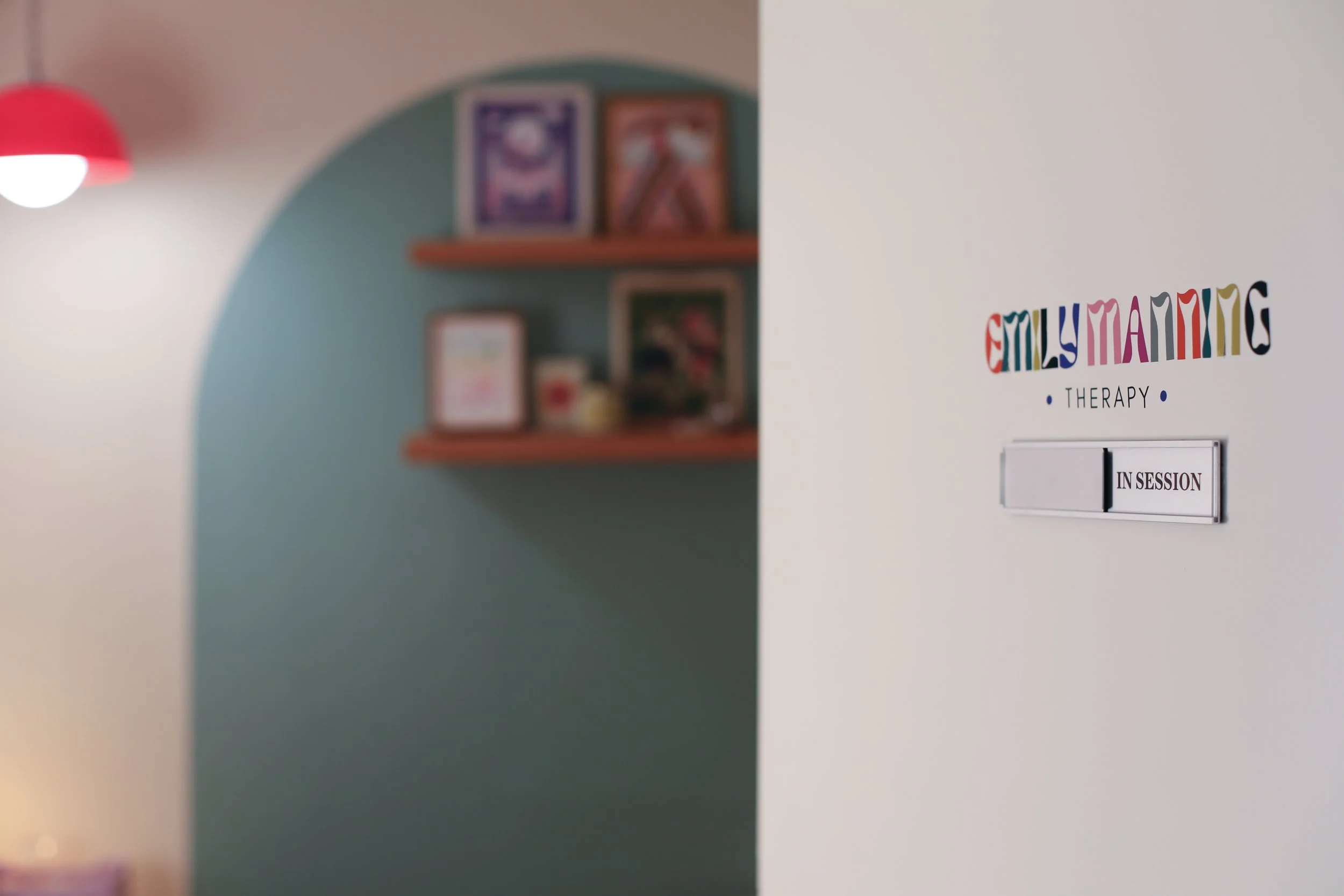 Partial view of a therapy room with wooden shelves and queer-affirming artwork in the background, and a door sign that reads "Emily Manning Therapy."