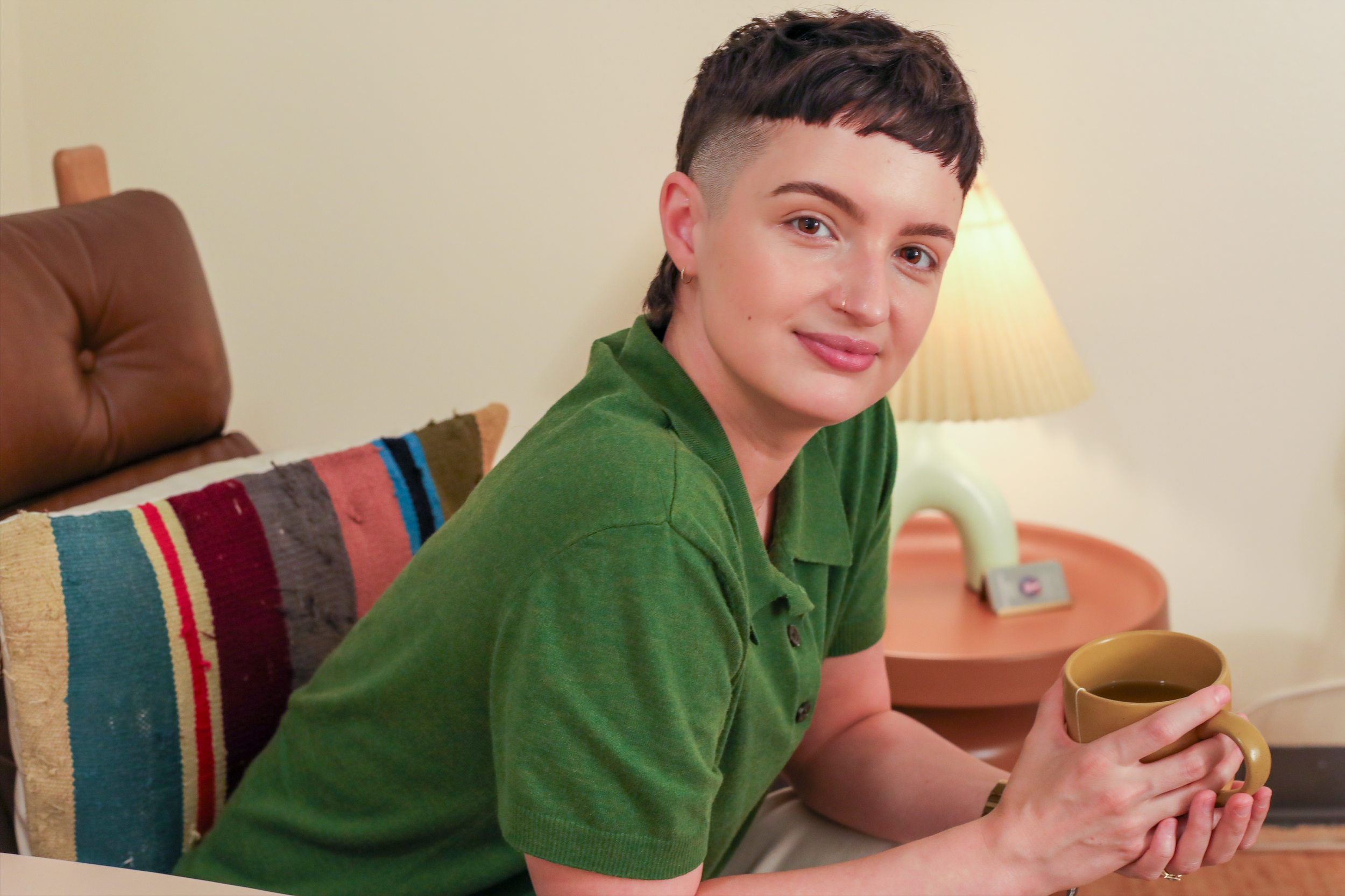 Queer therapist with short dark hair and an earring sitting in a brown leather armchair, holding a tea mug, in a cozy therapy room with a lamp and a colorful striped pillow.
