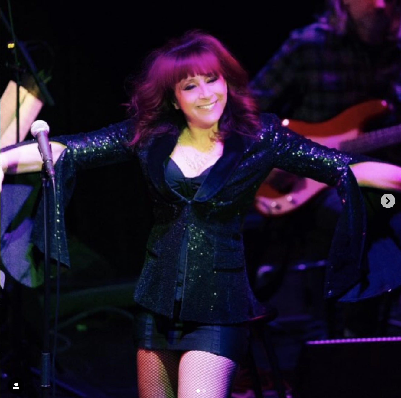Performer on stage smiling, wearing a black sequined jacket, black dress, fishnet stockings, and a necklace, with a microphone and guitar in the background.