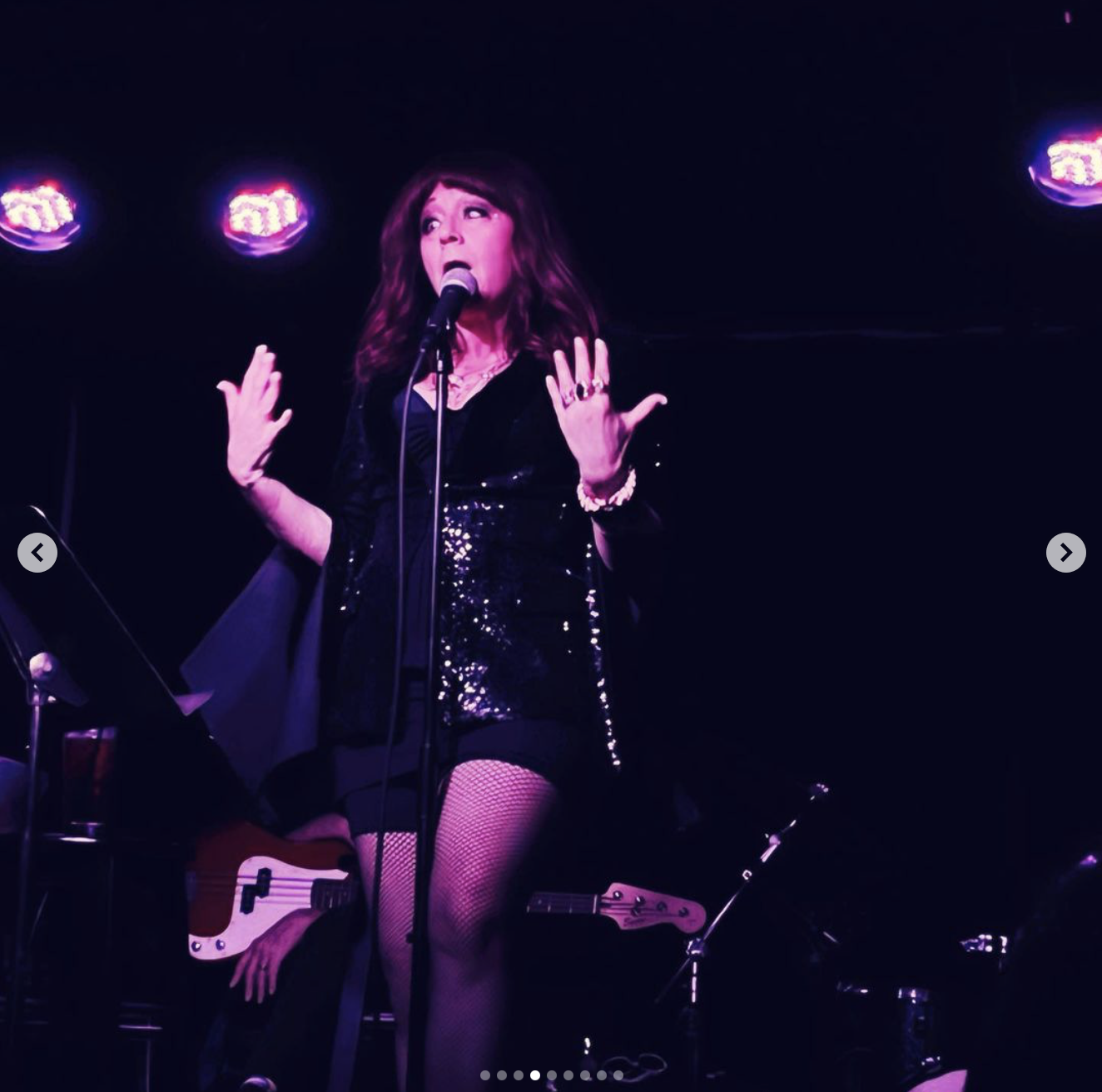 A woman singing on stage at a concert, wearing a black sequined dress, fishnet stockings, and jewelry, with purple stage lighting in the background.