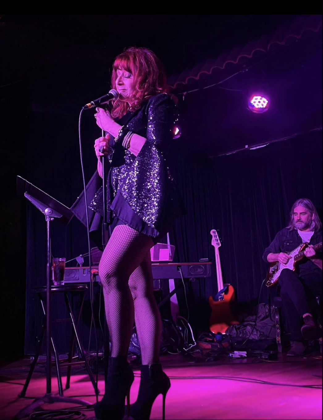 A woman performing on stage in a sparkly black jacket, fishnet stockings, and high heels, singing into a microphone, with a guitarist seated behind her.