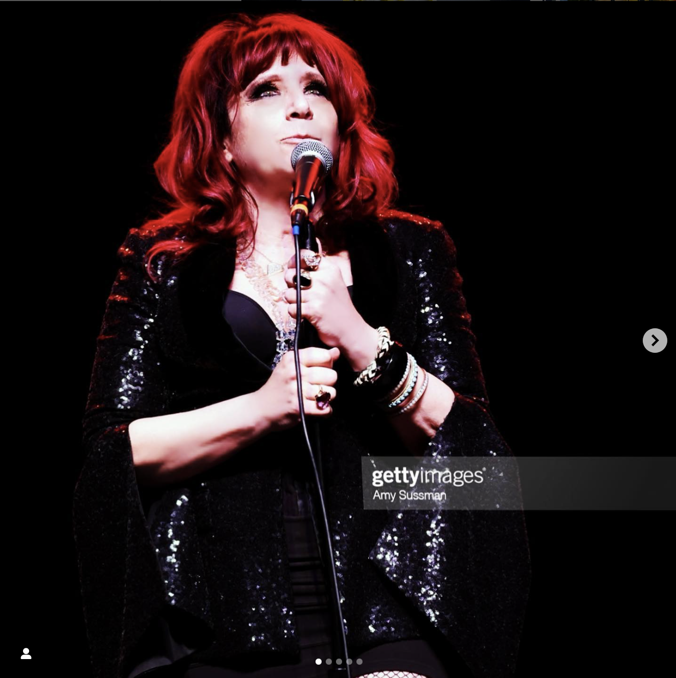 A woman with red hair in a black sequined jacket holding a microphone, performing on stage against a black background.