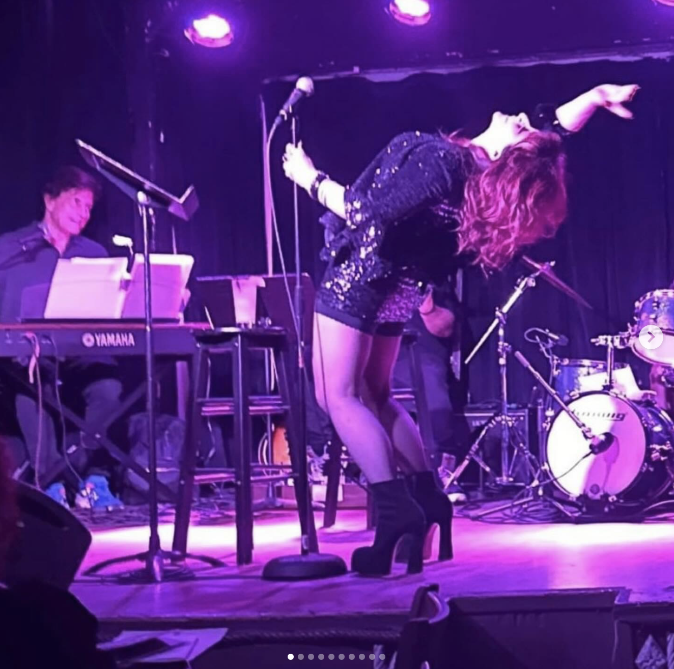 A woman with curly red hair dancing on stage during a musical performance, accompanied by a band including a keyboard player and a drummer, all under purple stage lights.
