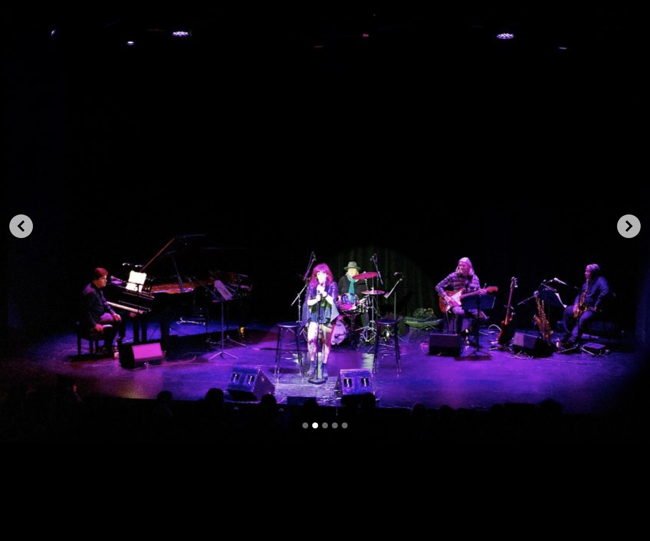 Live music performance on stage with six musicians, including a female singer, pianist, drummer, guitarist, saxophonist, and bassist, under purple stage lighting.