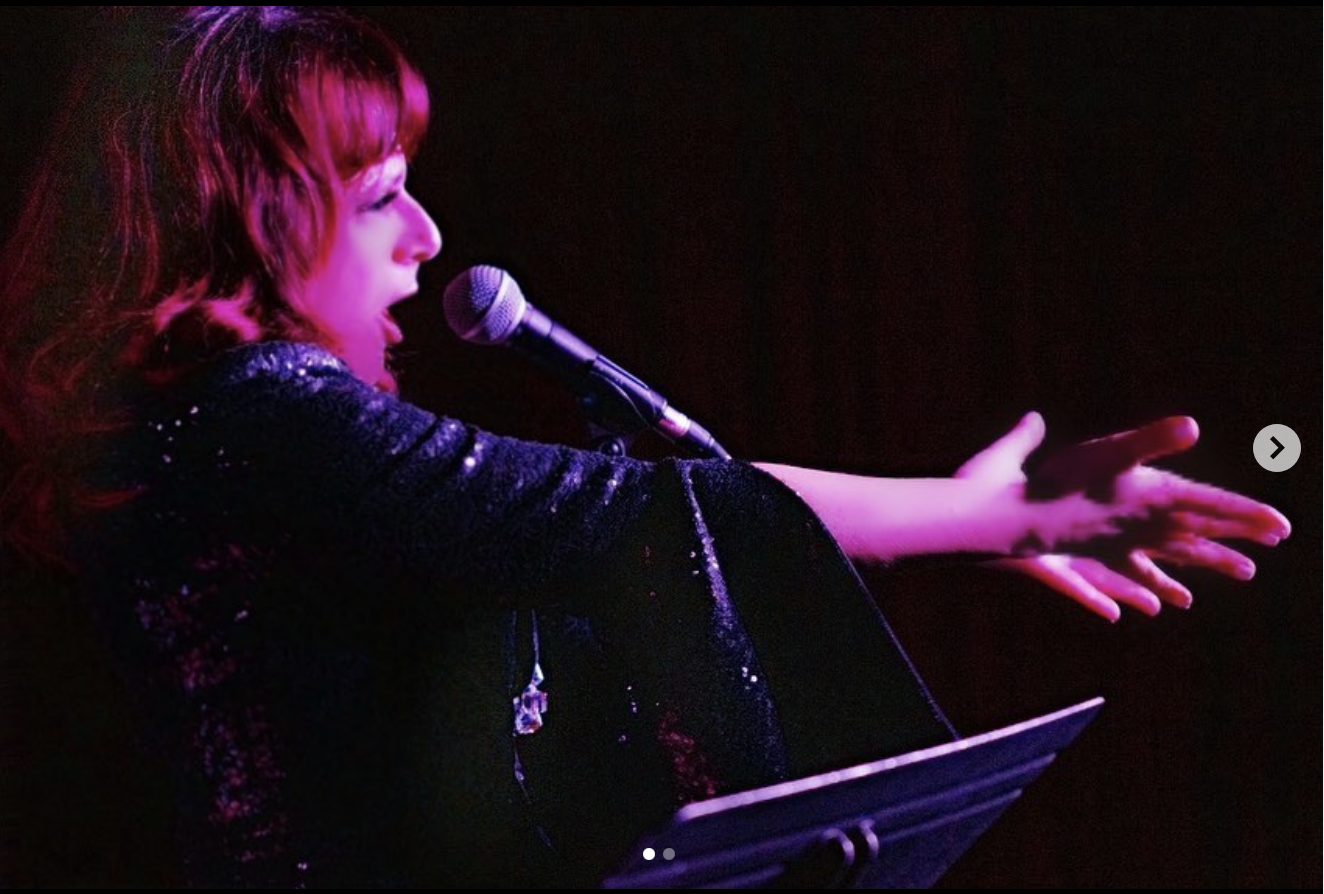 Performer with red hair singing and extending her hand on stage with a microphone