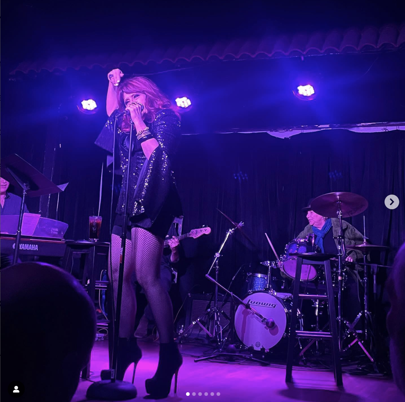 A female singer with long hair in a black sequined outfit, fishnet stockings, and high heels performing on stage, holding a microphone with purple stage lighting. A drummer and guitarist are in the background, playing their instruments.