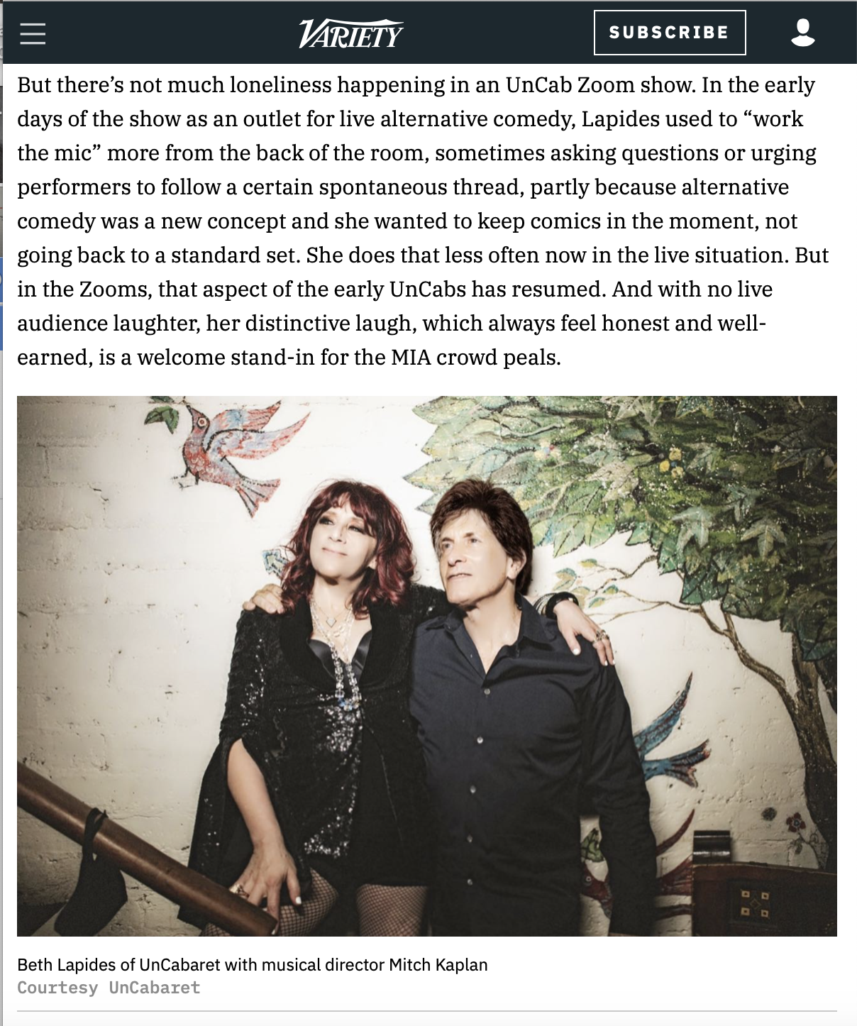 Beth Lapides and Mitch Kaplan posing together in front of a painted wall with a bird and trees, at a UnCabaret event.
