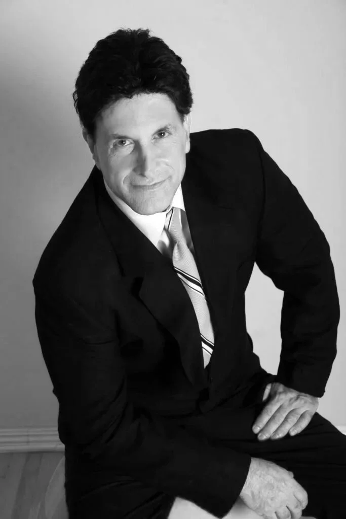 A man with dark hair, wearing a black suit, white shirt, and striped tie, sitting on a red stool against a plain white wall.
