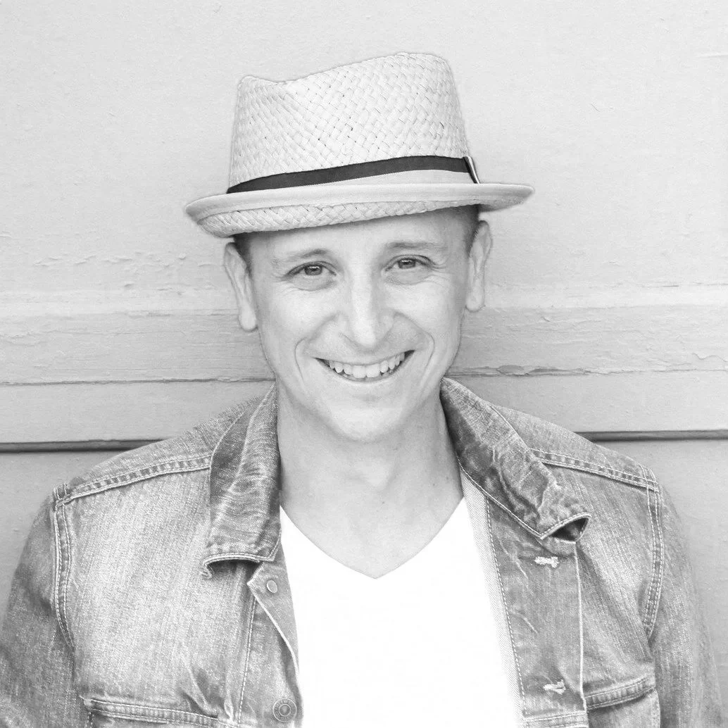 Man smiling, wearing a pink shirt and denim jacket against a blue background.