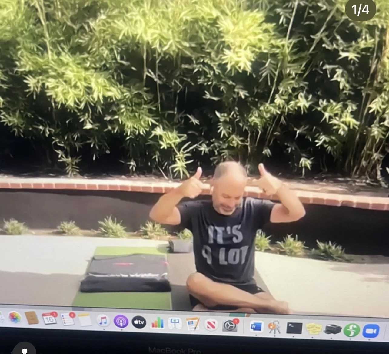 A person with a shaved head sitting cross-legged outdoors, smiling, and making thumbs-up gestures with both hands. The individual is wearing a black T-shirt with white text and is surrounded by green plants and a mats on the ground.