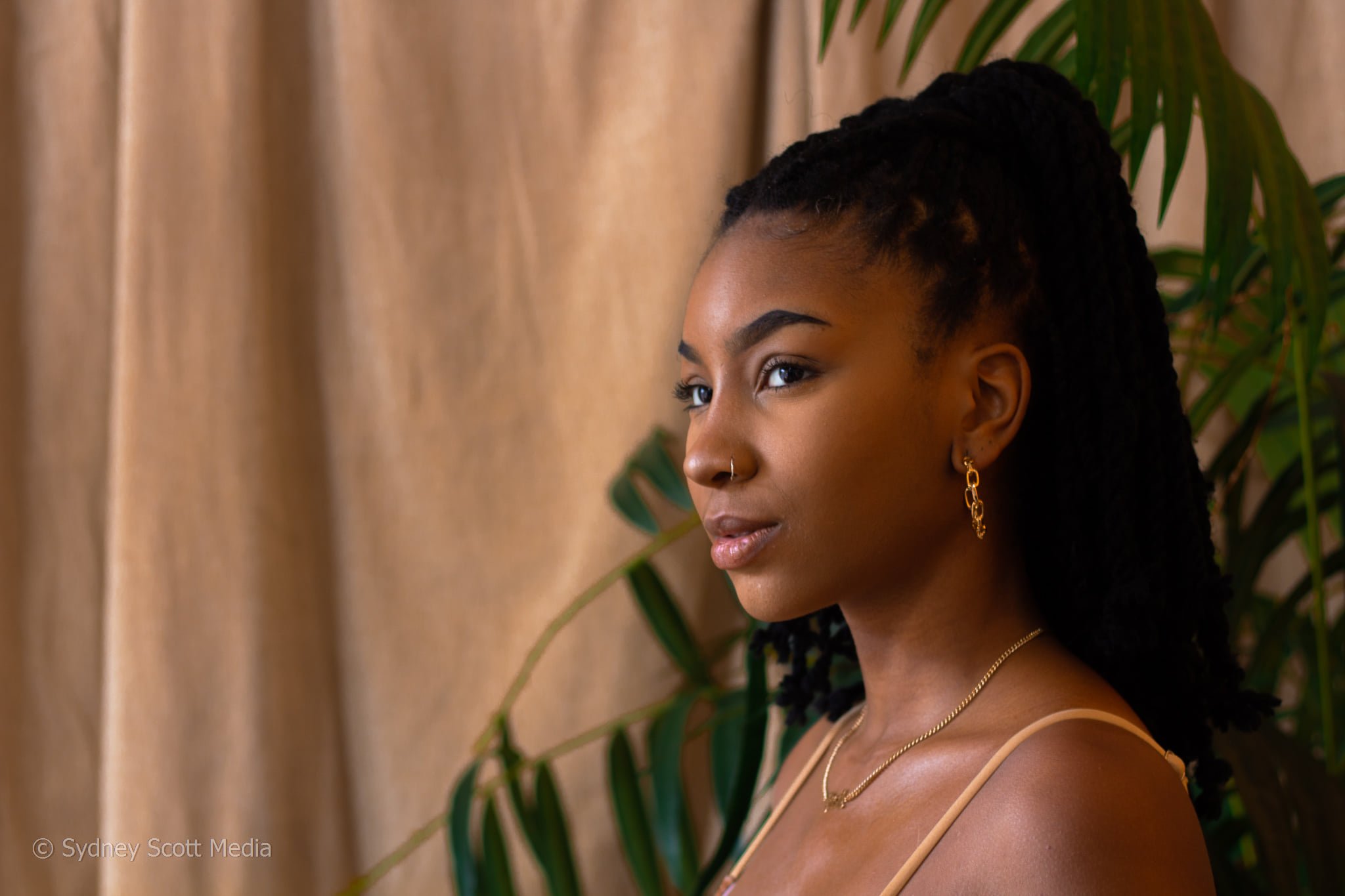 Profile of a young woman with dark braided hair, wearing gold jewelry, standing against a background with green leaves and beige curtains.