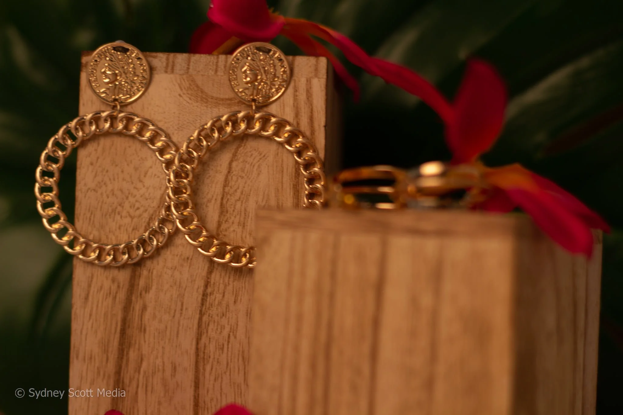 Gold jewelry, including a chain and earrings, displayed on a wooden stand with a red ribbon and green leaves in the background.