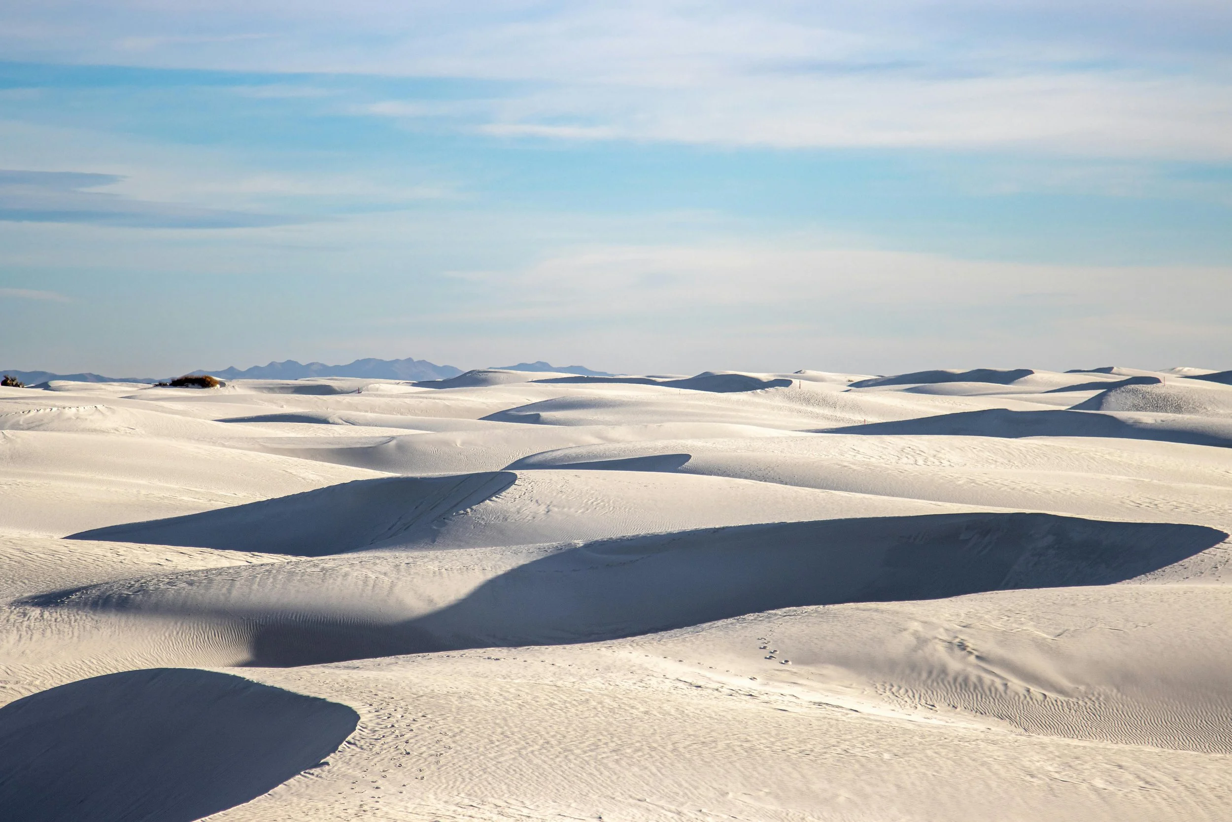 Snow-covered desert landscape with rippled snow dunes under a partly cloudy sky and distant mountain range.