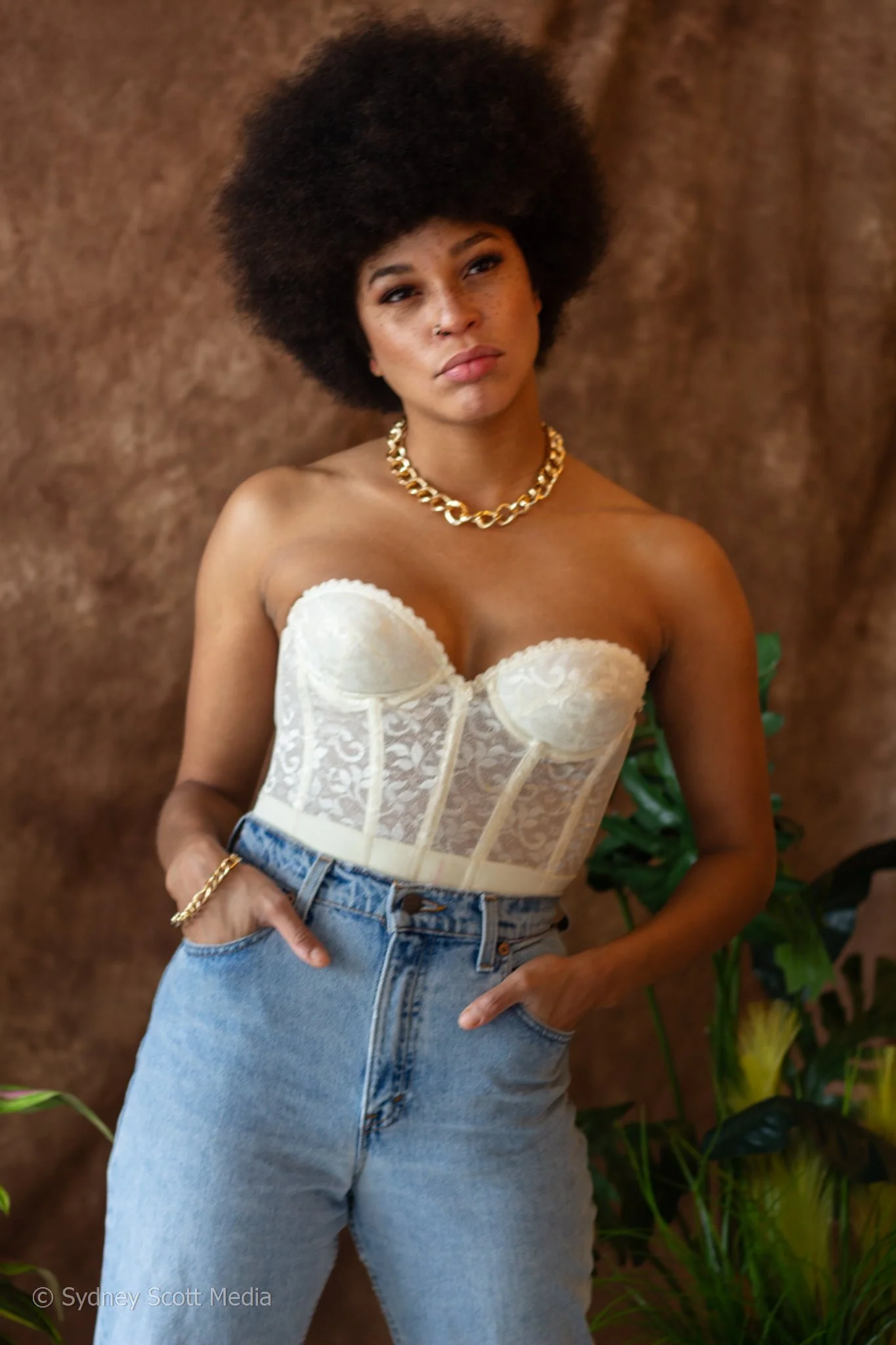 A woman with dark skin, freckled face, and full lips, poses with her right hand in her pocket, wearing a white, strapless lace corset top, light blue high-waisted jeans, a gold chain necklace, and a matching gold bracelet, standing against a brown textured background with green plants visible on the side.