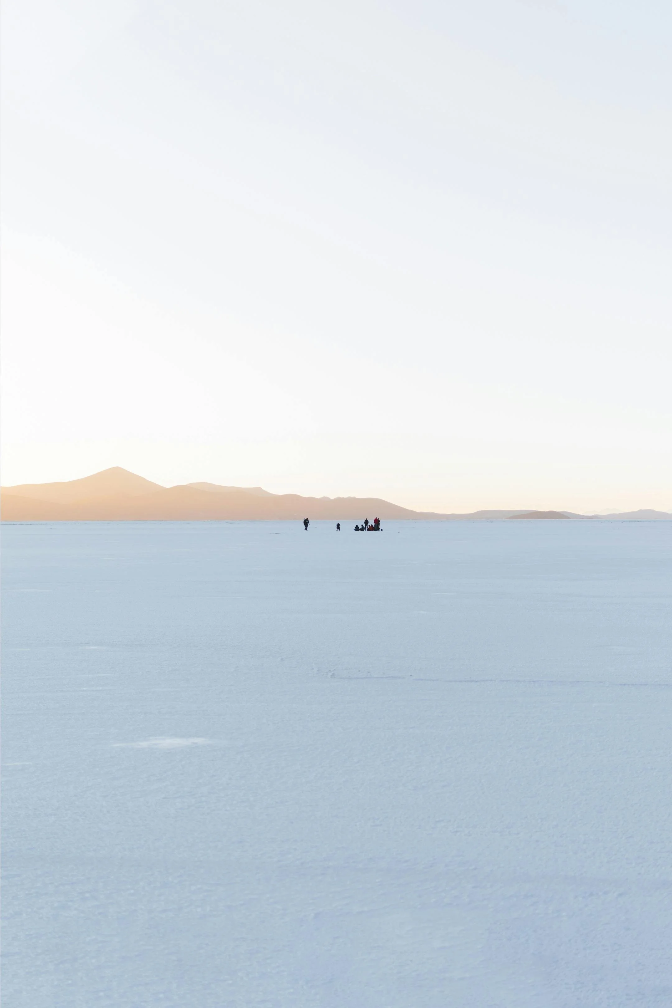 People walking across a vast, flat icy landscape with mountain ranges in the background, under a clear, pale sky.