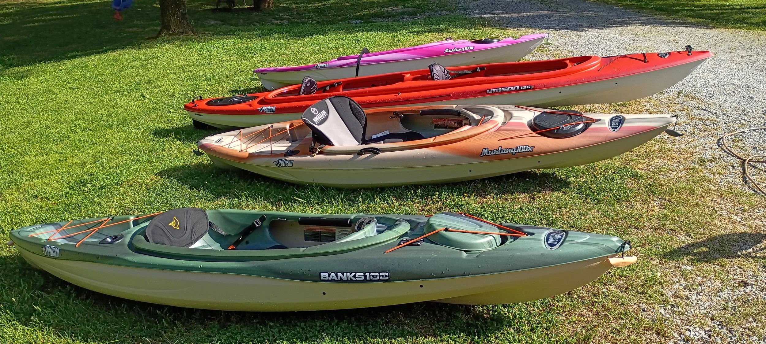 Four kayaks are lined up on grass and gravel, in green, peach, red, and pink colors with various gear inside.