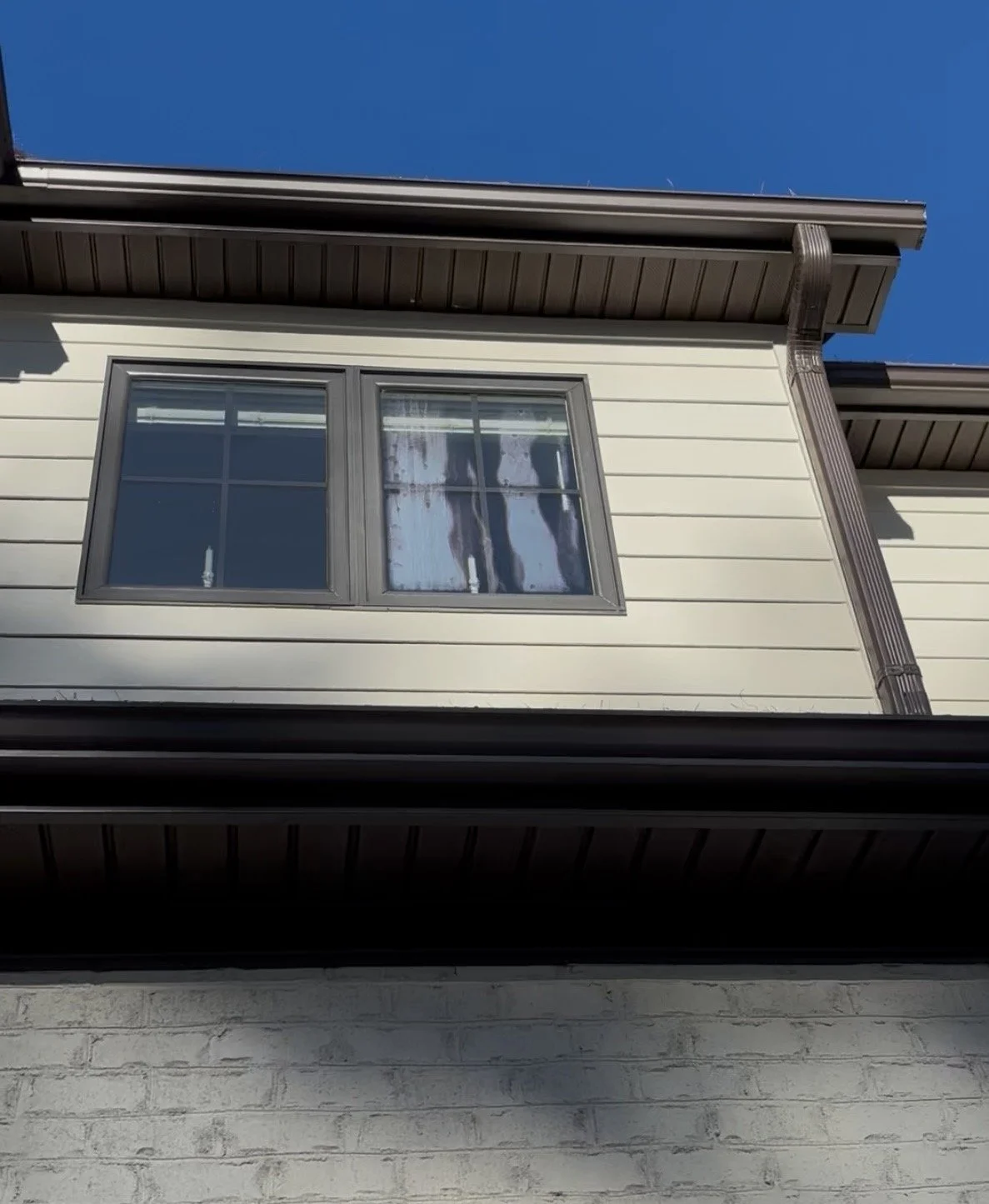 Close-up of a home's exterior showing a double window with gray trim, part of the beige siding, and a brown gutter against a clear blue sky.