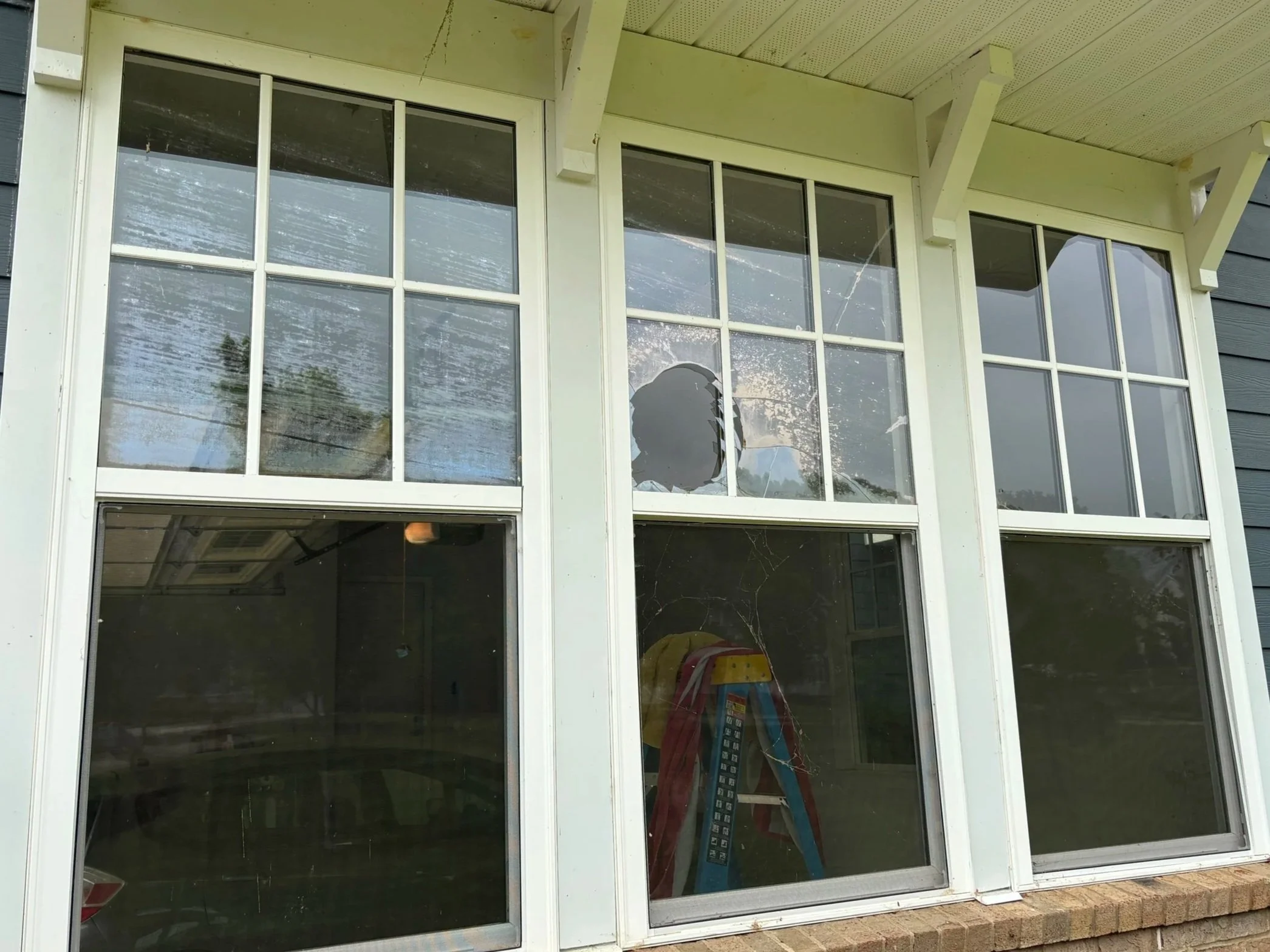 The image shows a large sliding glass door with multiple panes, some of which are broken. The door is part of a house with a light-colored exterior and a small brick foundation. Inside, a ladder and some clutter are visible.