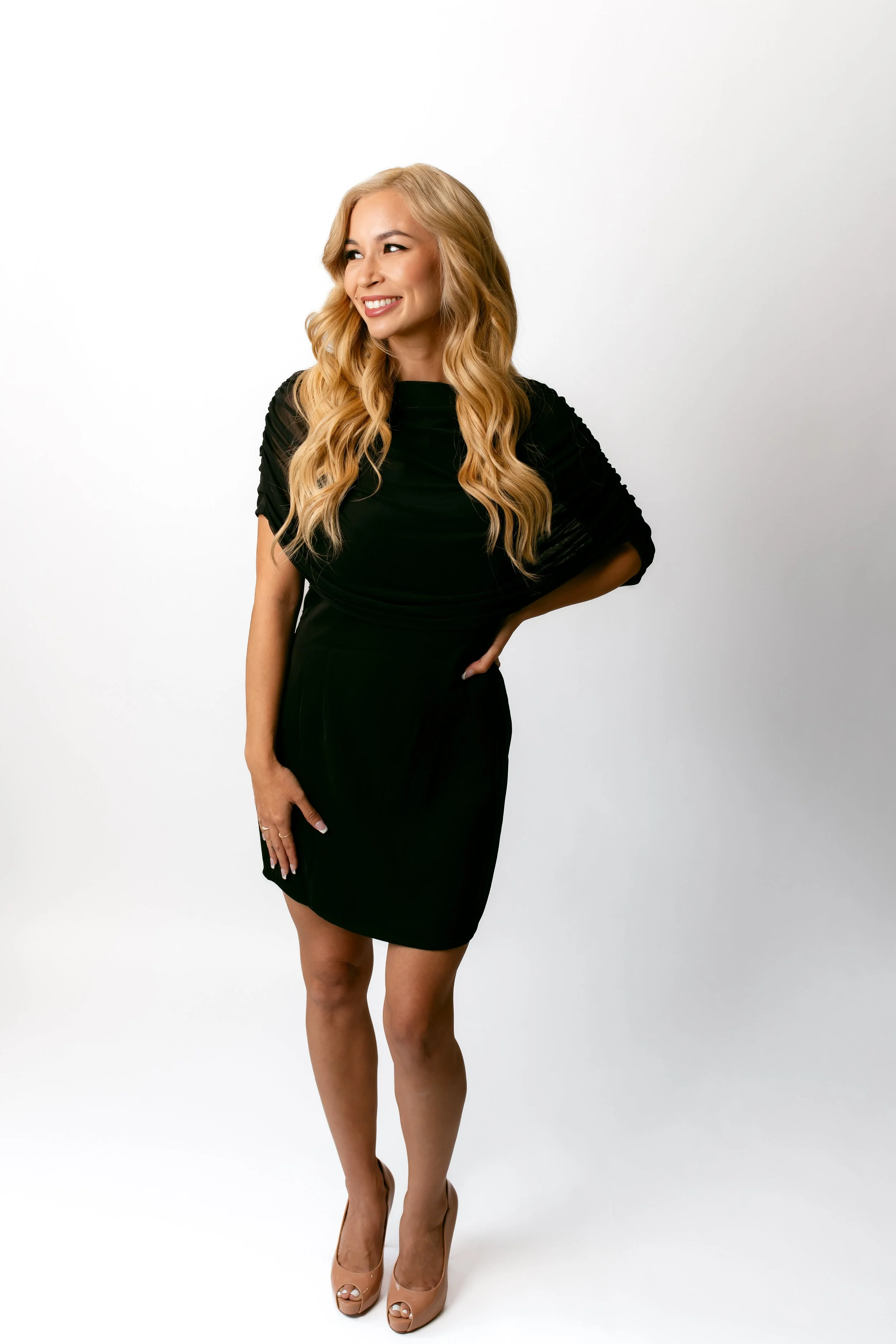 A woman with long wavy blonde hair wearing a black dress and nude high heels standing against a plain white background.