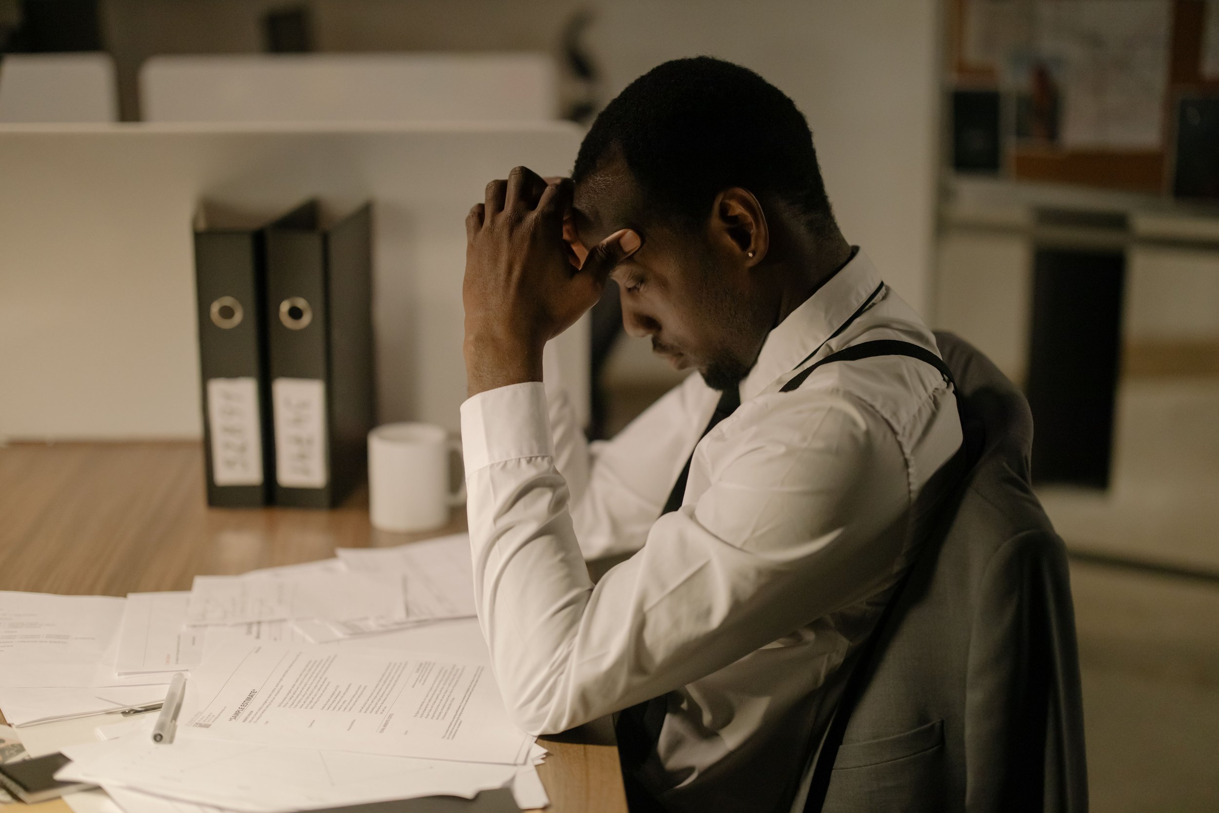 A man with short black hair, wearing a white shirt and black suspenders, sits at a desk with paperwork and a pen, resting his head in his hand in a dimly lit office space.