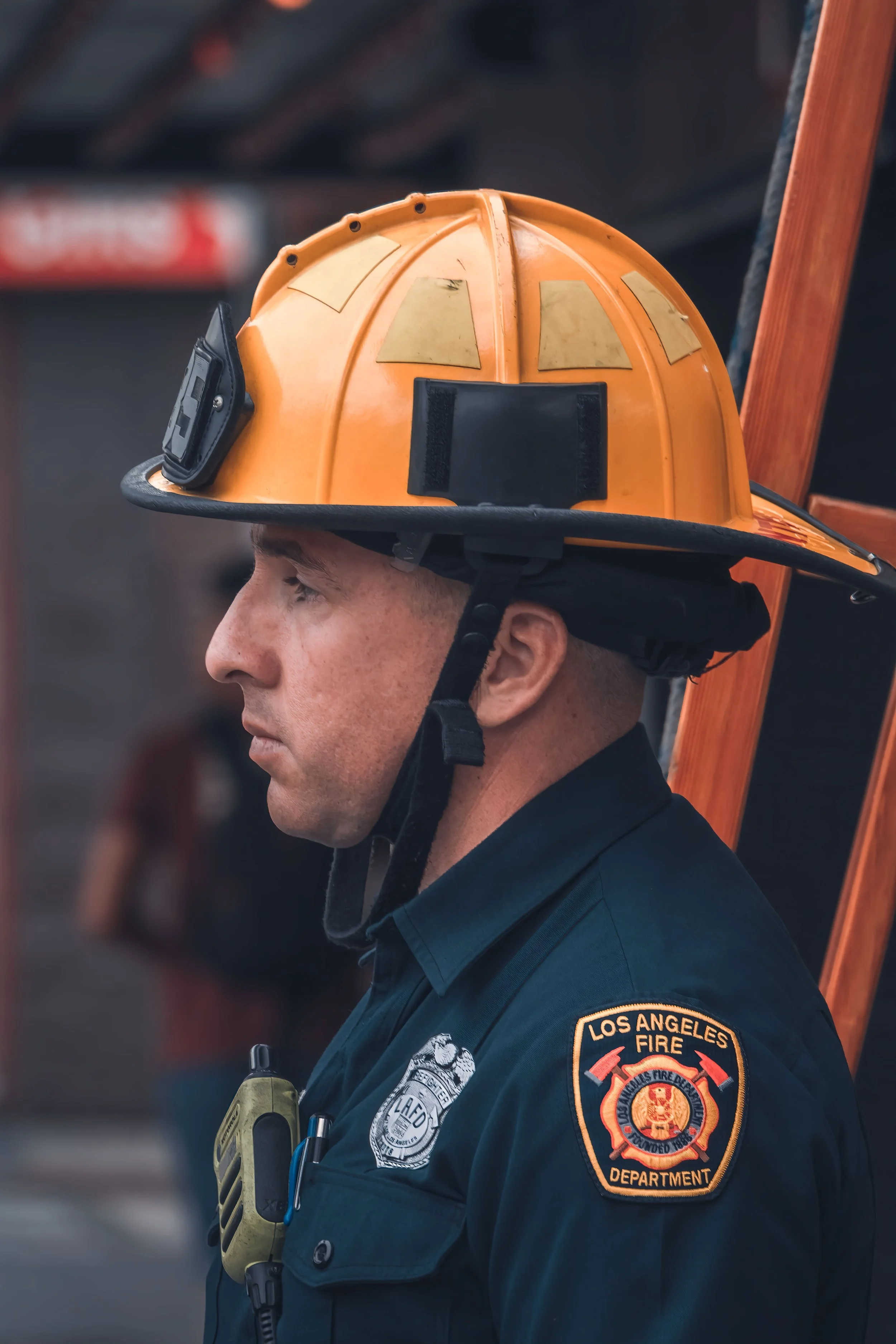 Profile of a firefighter in a navy uniform with a Los Angeles Fire Department patch, wearing an orange helmet with yellow reflective patches, and equipment attached to the uniform.