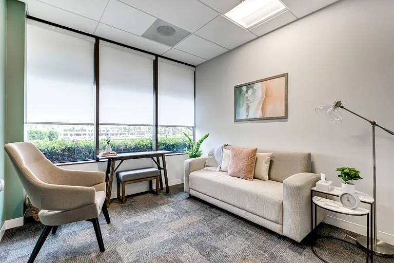 Modern office lounge with a beige sofa, armchair, window with blinds, side table, artwork, potted plant, and floor lamp.
