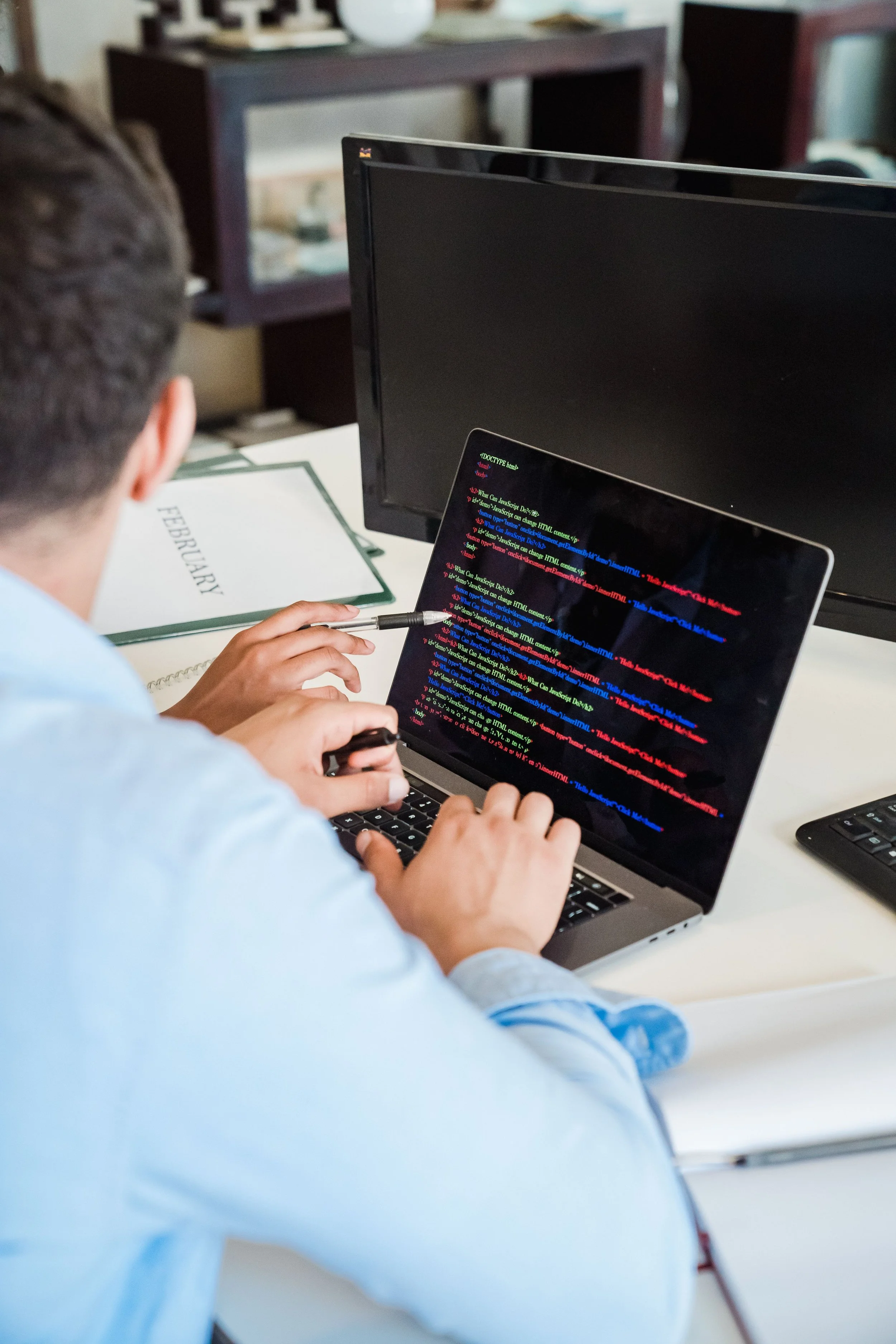 A person working on a laptop with programming code displayed on the screen, in an office setting with a calendar marked February and a computer monitor in the background.