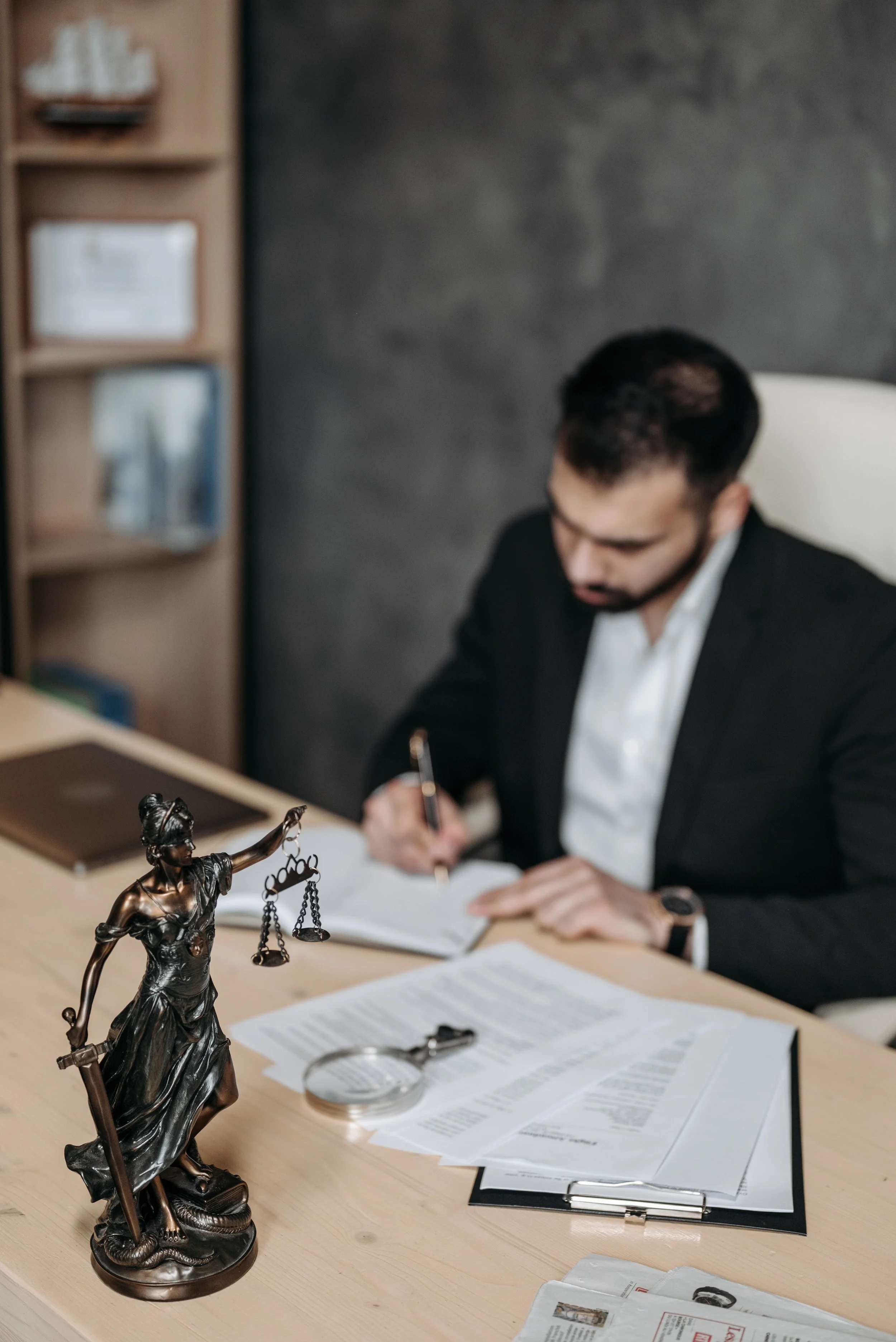 A man in a black suit and white shirt sitting at a desk, writing in a notebook with documents and papers scattered around, with a bronze Lady Justice statue in the foreground on the desk.