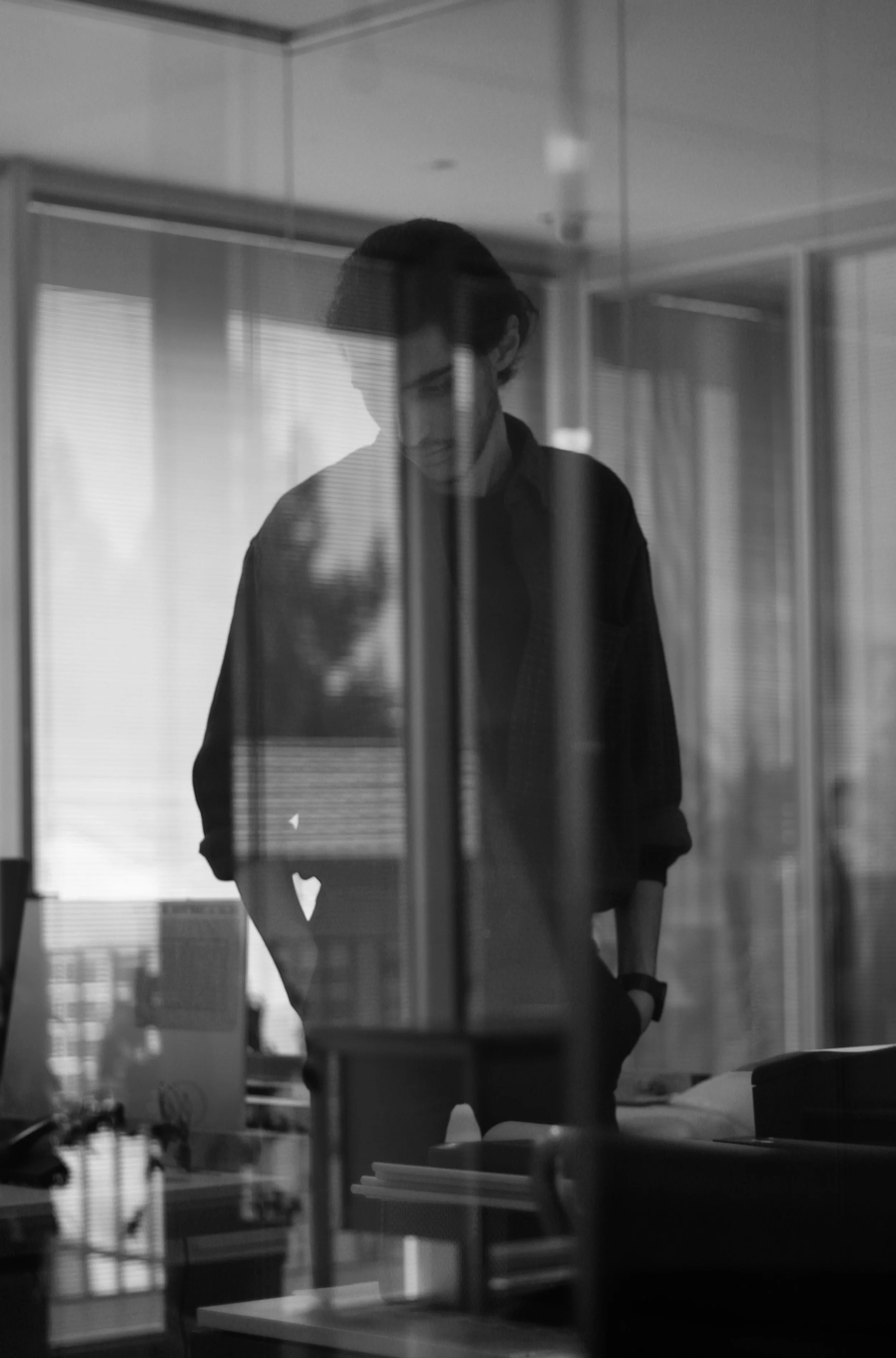 A black and white photo of a man with dark hair standing behind glass, looking down, with desks and office supplies in the background.