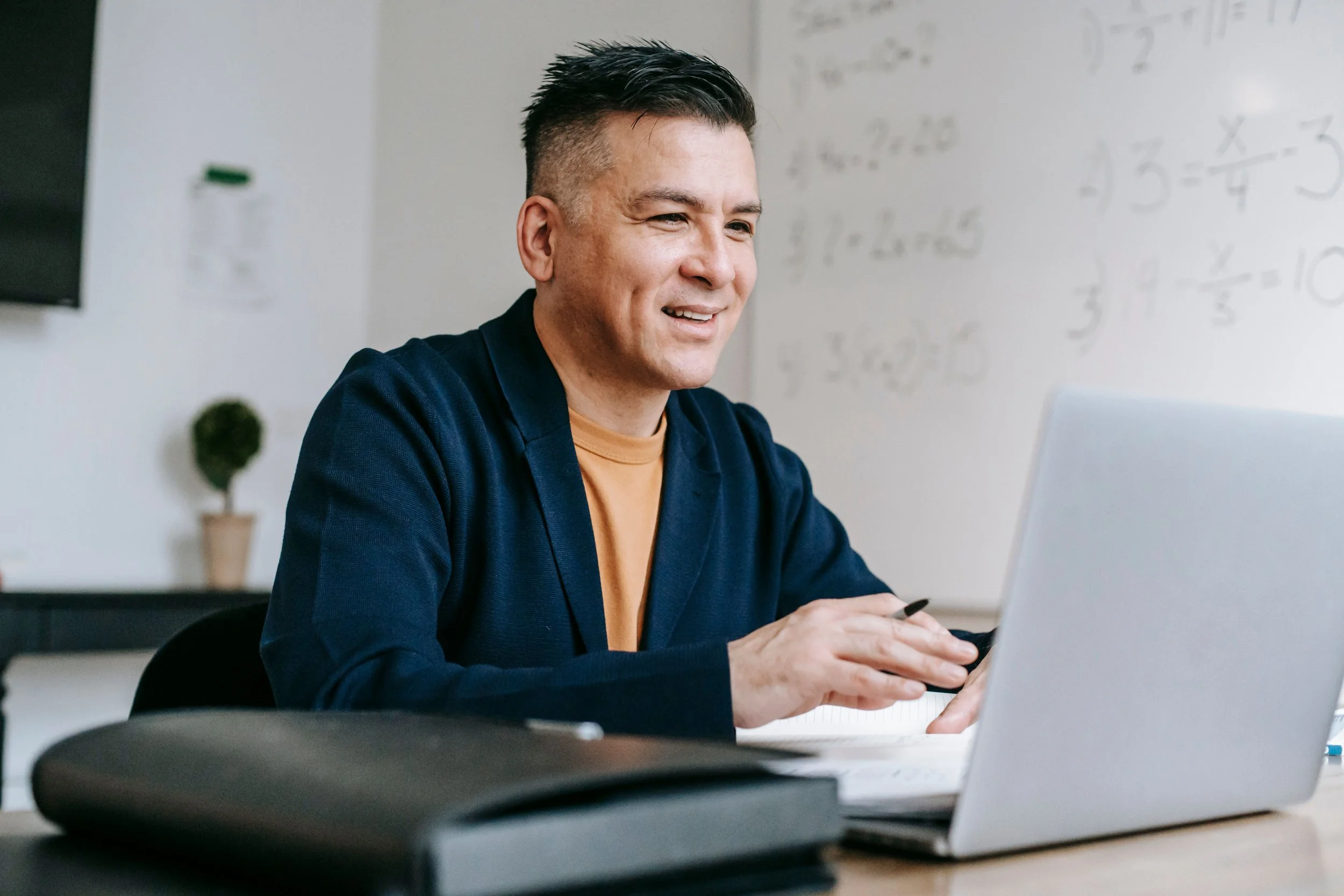 A young man with dark spiked hair and light skin, smiling and looking at a laptop computer in an indoor setting, with a whiteboard full of numbers and calculations in the background.