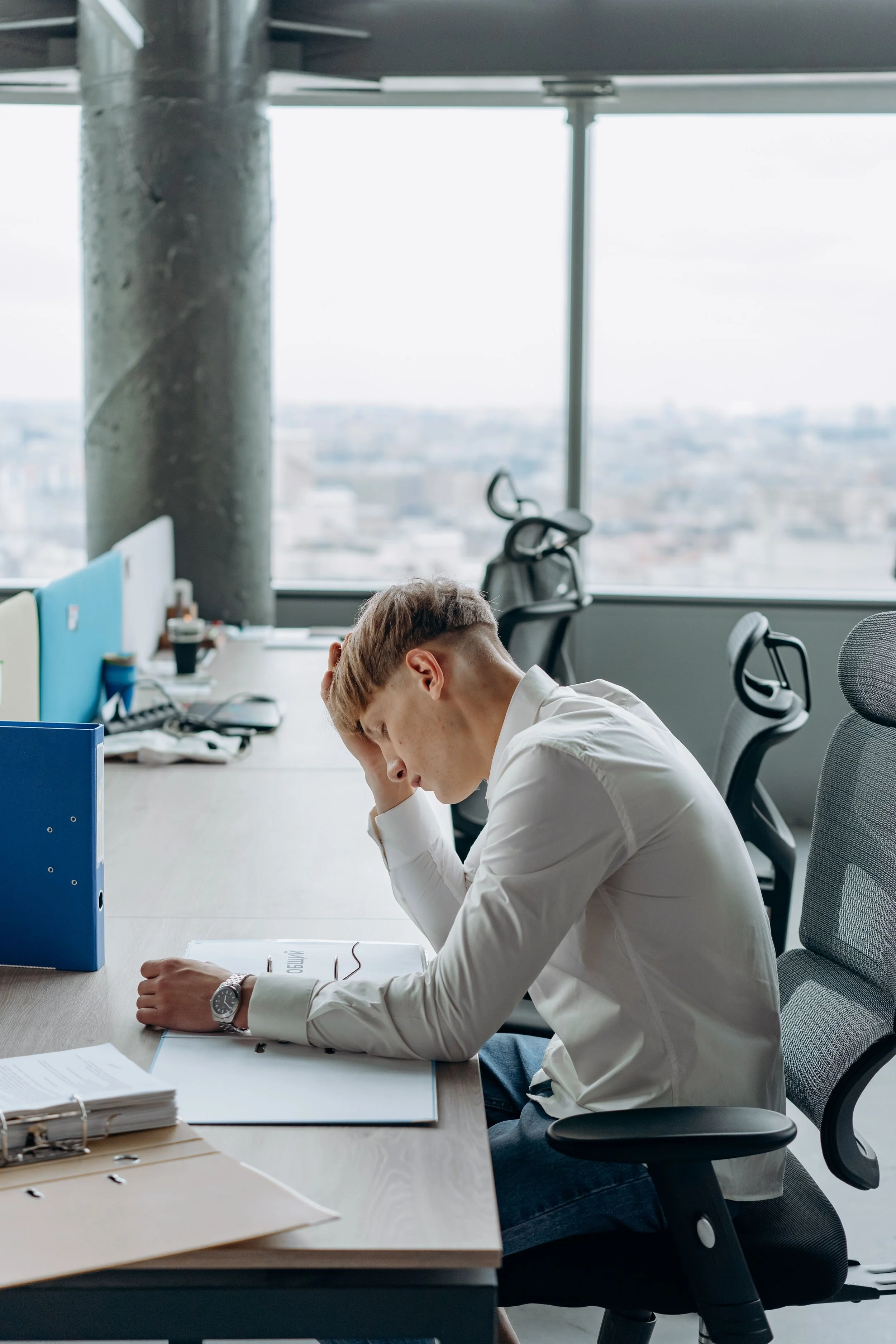 An office worker in a white shirt sitting at his desk, appearing stressed or overwhelmed, with papers and a binder in front of him, in a modern office space with large windows and city view.