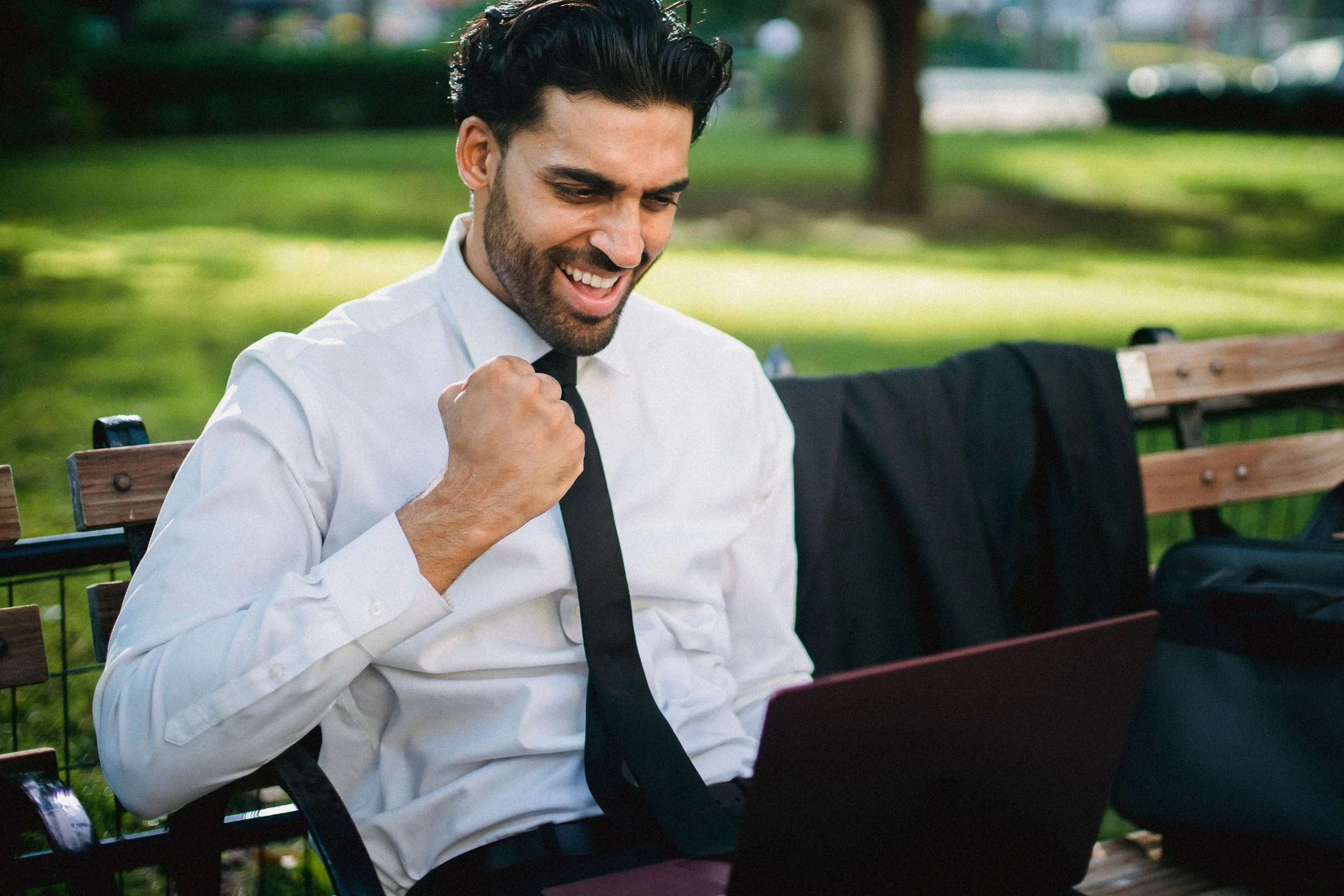 A man in a white shirt and black tie sitting on a park bench, smiling and clenched fist in celebration, with a laptop on his lap, outdoor park setting in the background.