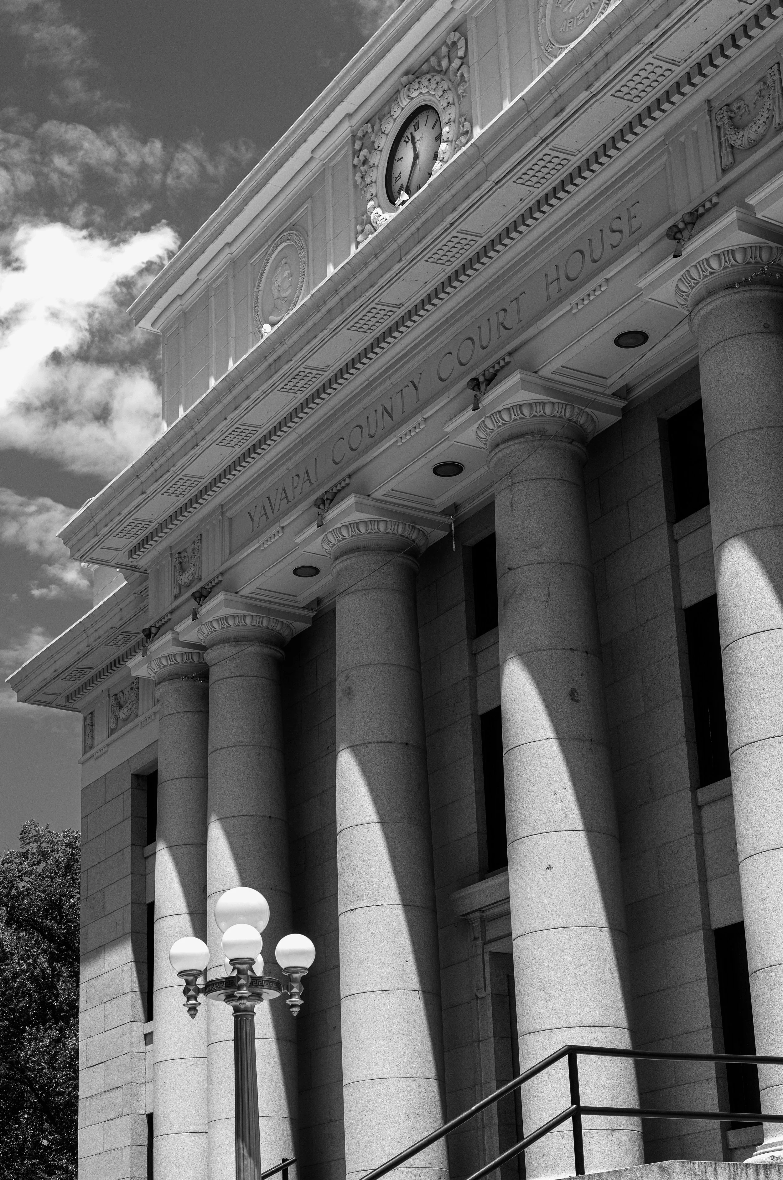 The exterior of the Yavapai County Courthouse featuring large stone columns, a decorative clock above the entrance, and a lamppost in front. The photo is in black and white.