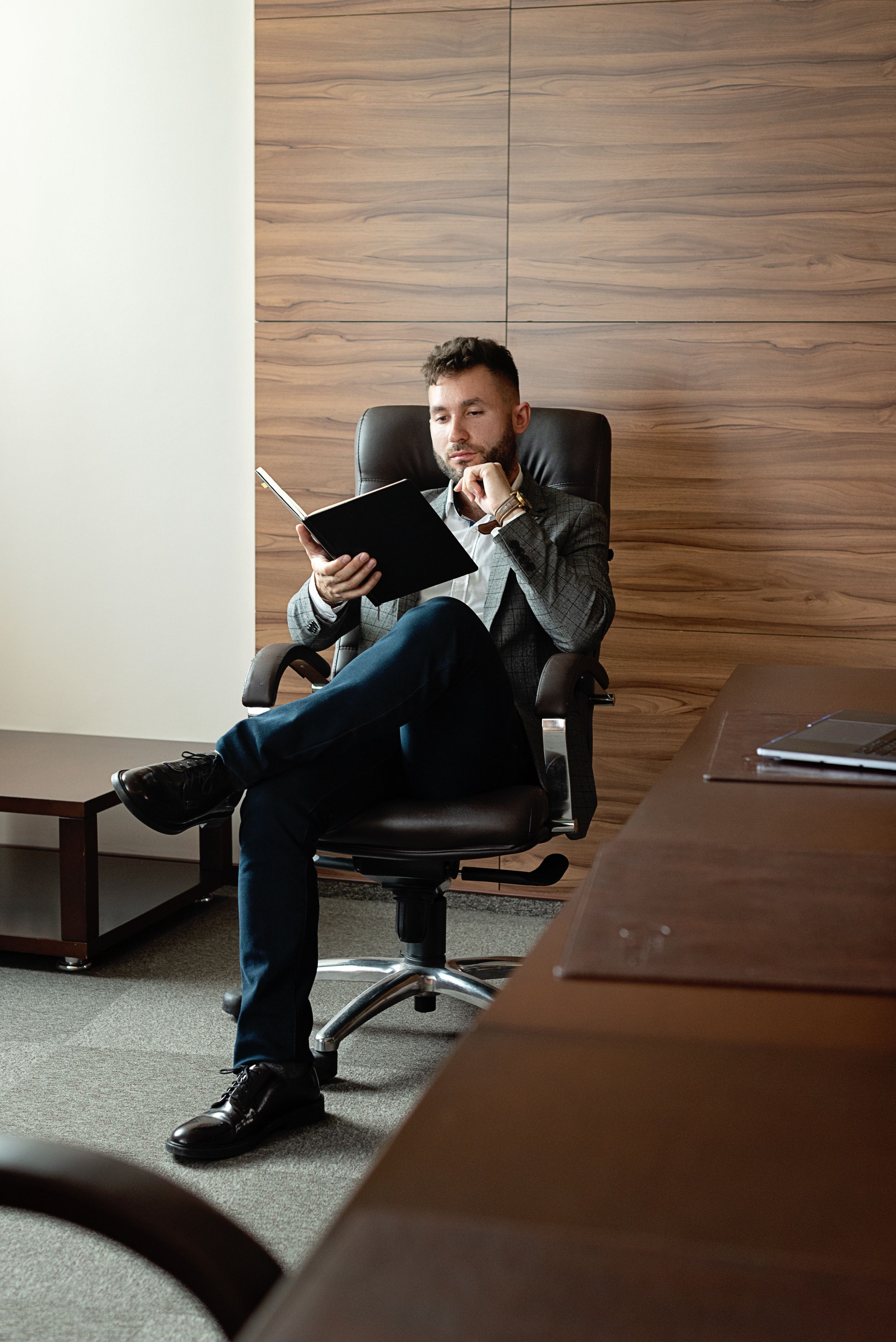 A man in business attire sitting in an office chair, reading a book, with a serious or thoughtful expression, in front of a wooden wall with a light-colored wall on the left.