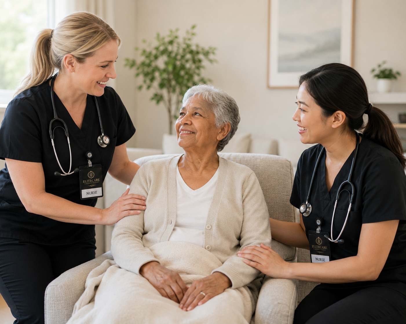 An elderly woman sitting in a beige armchair, smiling at two female nurses who are tending to her at home. The nurses are wearing black scrubs with stethoscopes around their necks and name tags that say 'NURSE'.