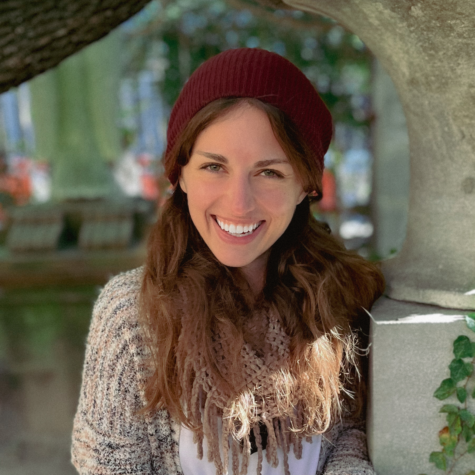 A young woman with red hair wearing a red beret, smiling, standing outdoors under a stone archway, with blurred greenery in the background.